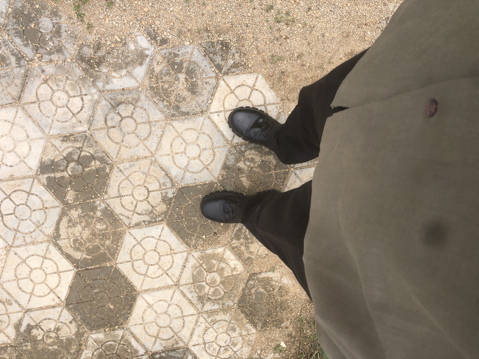 This is a first-person perspective media file showing a person's feet and lower legs standing on a patterned pavement. The pavement consists of hexagonal tiles, each with a circular design resembling a flower with petals. Some of the tiles are lighter in color, while others are darker and appear wet or stained. The ground around the pavement is covered with small pebbles and some sparse green vegetation.
The person is wearing dark trousers and black, sturdy boots with laces. The boots appear to be work boots or hiking boots. The fabric of the trousers is dark and matte. A portion of a light-colored garment, possibly a jacket or shirt, is also visible, with a dark button or emblem on it. The person's posture suggests they are standing still.
The lighting in the image is somewhat diffused, indicating it might be daytime with overcast skies or the sun is not directly overhead. There are no specific clues about the time of day, but the general brightness suggests it is not nighttime. The presence of what looks like moisture on the pavement could indicate recent rain or dew, suggesting a humid or recently wet environment. The background of pebbles and sparse vegetation could be found in many outdoor locations. The location context provided is Gwafan, Nigeria. No text is visible in the image.