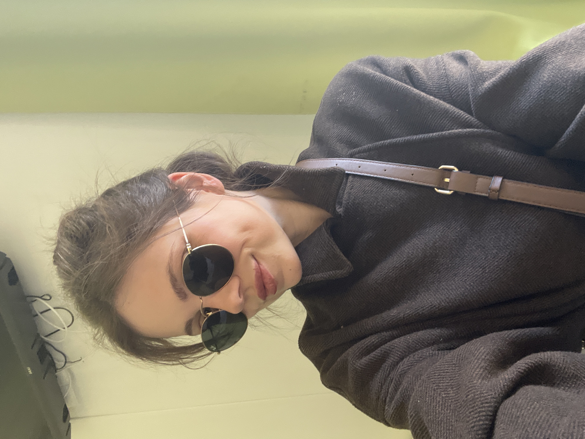 A woman is looking at the camera, wearing sunglasses and a dark coat with a brown strap across her chest. Her hair is pulled back. The background is plain and pale yellow, possibly a wall. The image has been rotated. No information in the picture suggests this is in Venice, Italy.