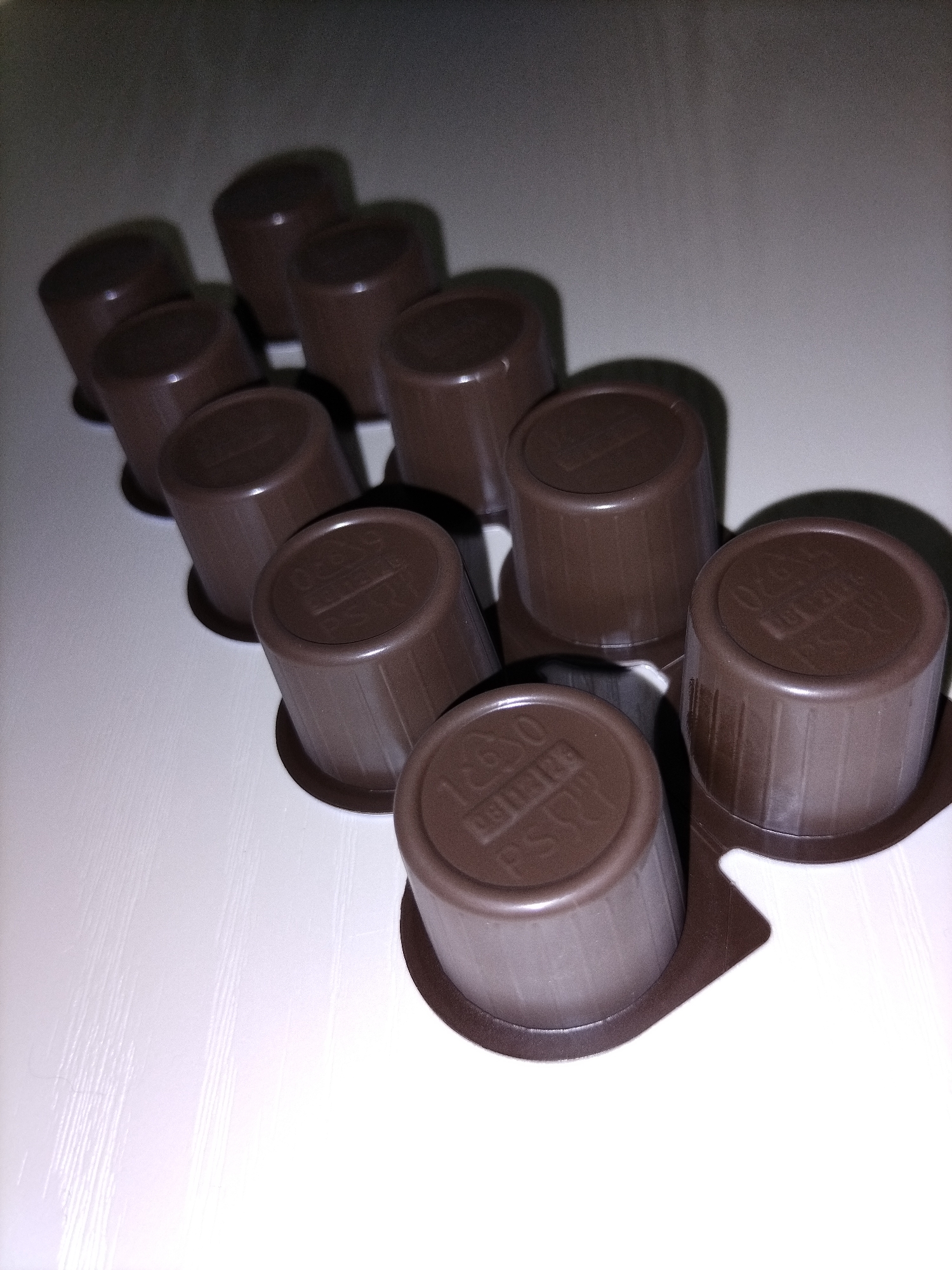 The image shows a group of small, brown, cylindrical containers, possibly coffee pods, arranged on a white surface. The containers are made of plastic and have ridged sides. Some of the containers have text and symbols embossed on their lids, but they are not clearly legible. The lighting in the image is dim and creates strong shadows. The background is a plain, dark surface, which is out of focus. There are no people or animals present in the image. The image does not provide any information about a specific location in the city of Смалявічы, Belarus.