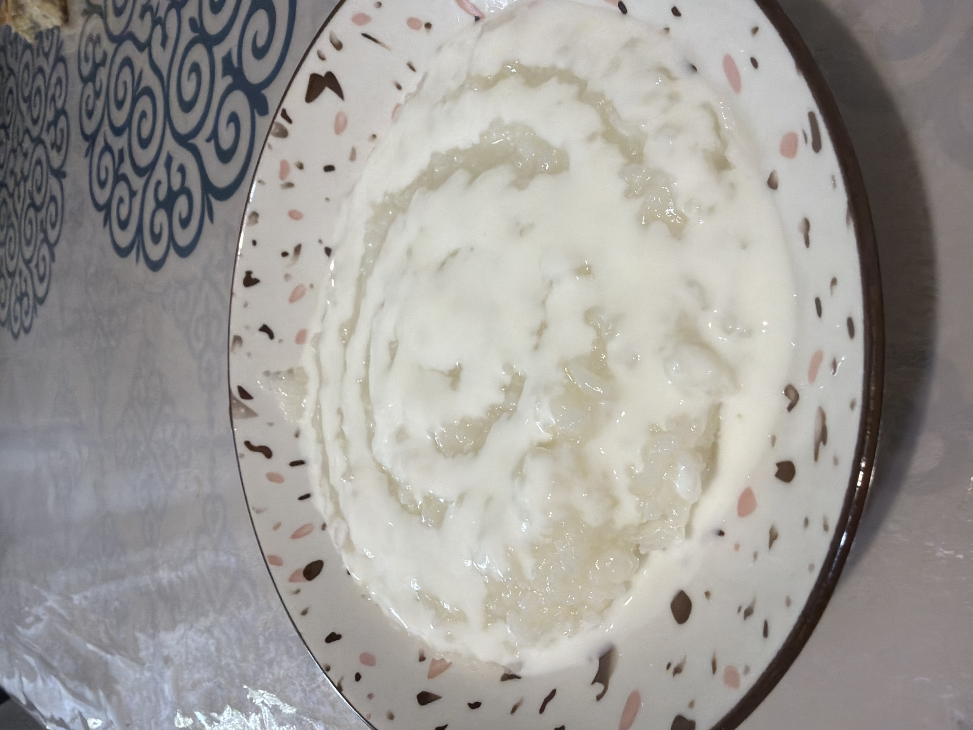 A close-up, overhead shot shows a white bowl with a brown rim and speckles of brown and pink, filled with a creamy white dessert. The dessert appears to be rice pudding, with visible grains of rice mixed into a smooth, thick liquid. Swirls of the creamy liquid have been added to the top, creating a decorative pattern. The bowl rests on a table covered with a light-colored, patterned tablecloth. The pattern on the tablecloth consists of swirling blue shapes. To the right of the bowl, a small piece of bread or pastry is visible. The lighting is soft, suggesting an indoor setting, possibly a dining room or kitchen. No people or actions are depicted, focusing solely on the food and its presentation. The image does not provide any specific visual cues to pinpoint its location within the city of Astana, Kazakhstan.