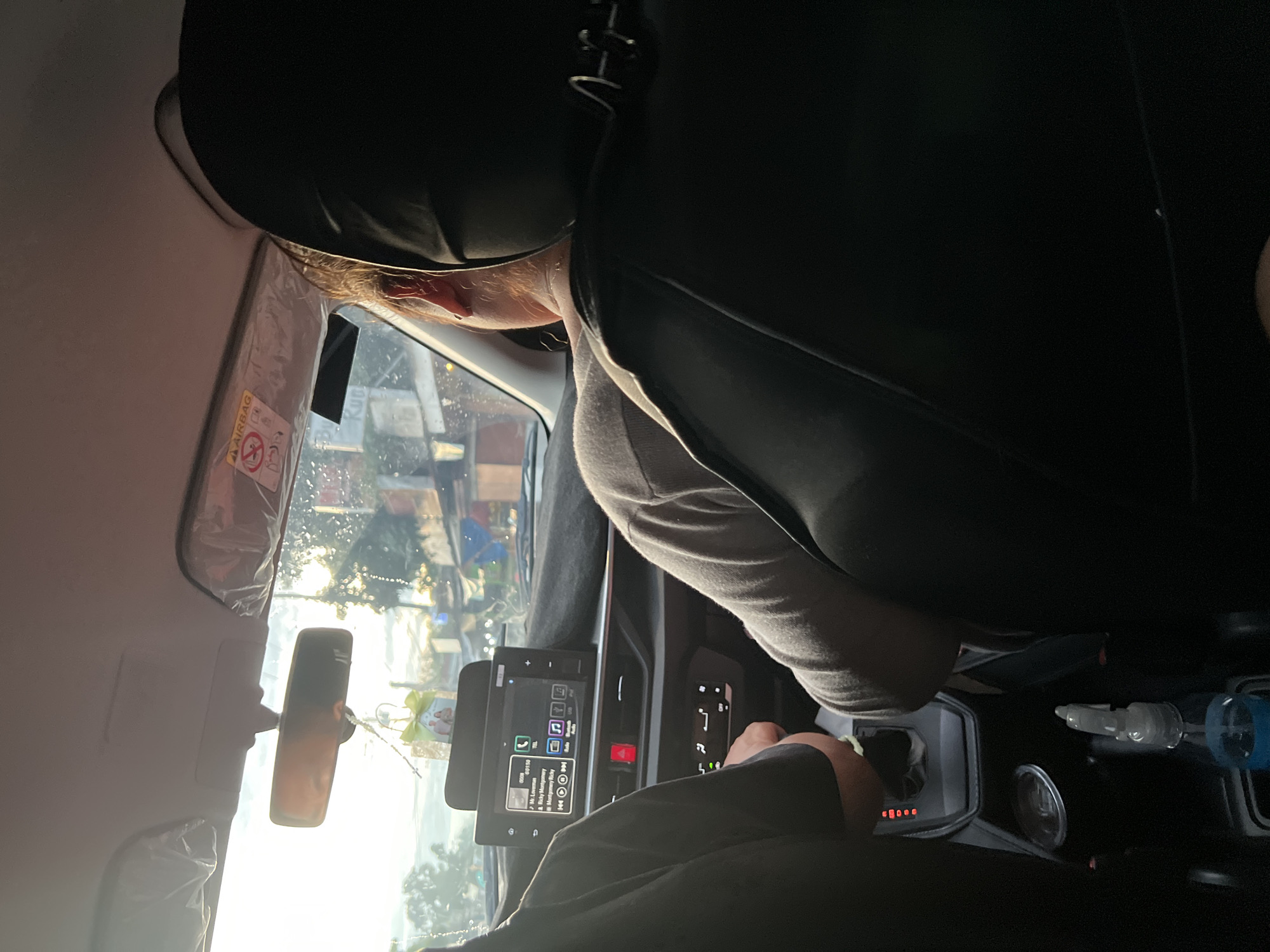 The image shows the interior of a car, likely taken from the passenger seat. The driver, a person with visible skin on their arms and a light-colored shirt, is operating the vehicle. The car's infotainment screen is visible, displaying music or navigation information. Through the windshield, a cityscape can be seen, and based on visual clues, it is strongly indicated this is Davao City, Philippines. This is suggested by the type of buildings and streetscape visible through the windshield. The sun visor on the driver's side is partially extended. Items such as a hand sanitizer and what appears to be a drink container are present in the center console. The perspective is from the back seat, looking forward towards the driver and the dashboard.