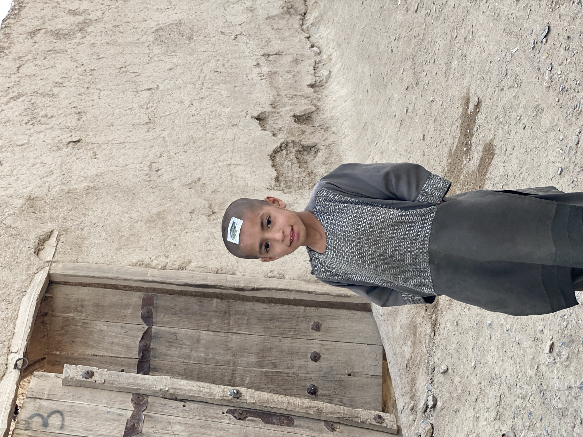 A young boy with a shaved head stands in front of a weathered wooden door. He is wearing a dark gray tunic with a patterned bib. A small sticker, possibly a temporary tattoo, is affixed to the top of his head. The boy is looking at the camera with a slight smile. The background consists of a rough, textured wall made of mud or plaster, and the ground is covered in dirt and small stones. The scene appears to be outdoors, likely in a village setting, given the rustic architecture and the arid ground. The lighting suggests it is daytime, possibly a sunny day due to the shadows, although the overall mood is muted. The image captures a candid moment of the boy. There is no visible text in the image.