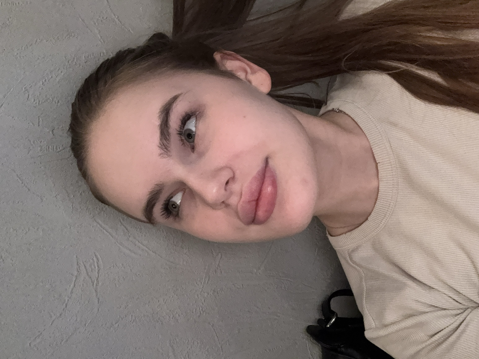 This is a close-up, vertically oriented selfie of a young woman, taken indoors. She has long, dark brown hair pulled back. Her face is turned to her left, and her gaze is directed to her left as well. She has full lips, parted slightly, and appears to be smiling subtly. Her eyebrows are well-defined, and her eyelashes are long and dark. She is wearing a light-colored, ribbed t-shirt. A thin silver chain is visible around her neck. The background is a textured, gray wall. Partially visible at the bottom right is a black bag or purse with a metal buckle. The lighting suggests it is daytime, possibly late afternoon or early evening, indoors with artificial light. There is no visible text in the image.