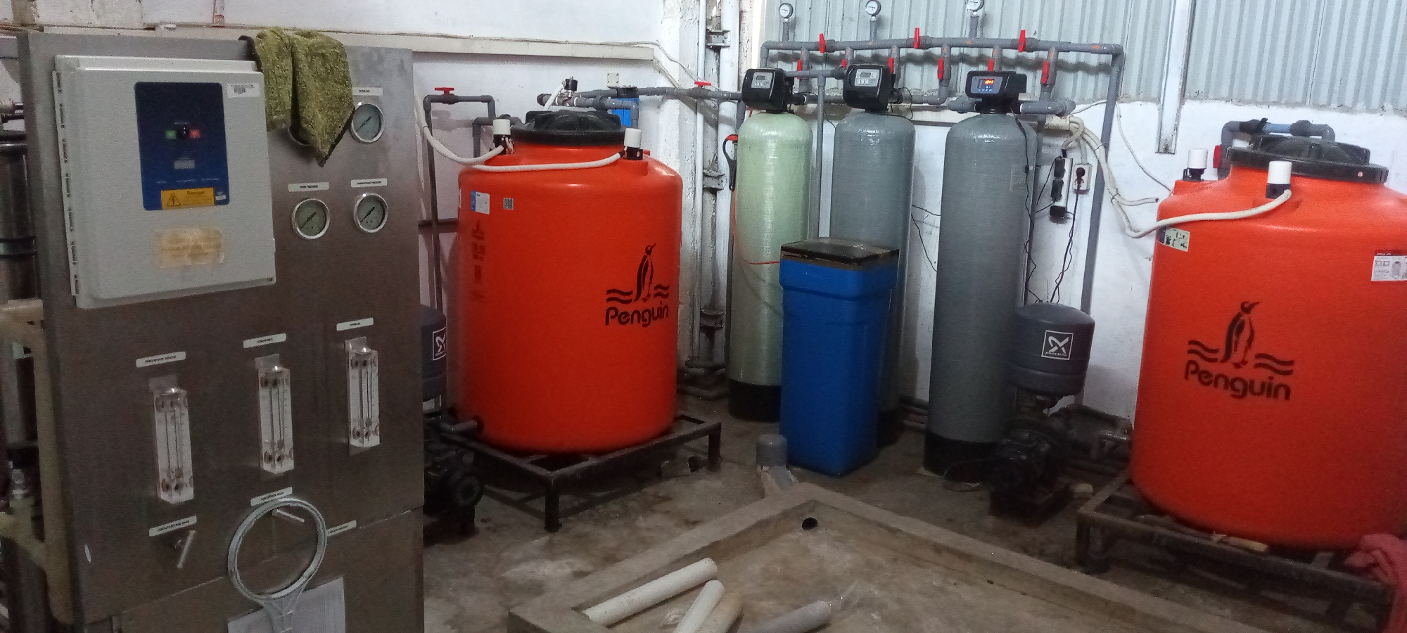 The image depicts a water purification system inside a room. There are two orange Penguin-branded water tanks on metal stands. Between them are three grey filtration columns with digital displays and red valve handles, a blue container, and a grey cylindrical container with an X logo. To the left, a stainless steel control panel features a digital display, pressure gauges, and flow meters. White PVC pipes are on the floor in front of a concrete basin. The location is likely in Lhokseumawe, Indonesia, given the branding of the water tanks are from a manufacturer in that location. The scene suggests an industrial or commercial setting for water treatment.