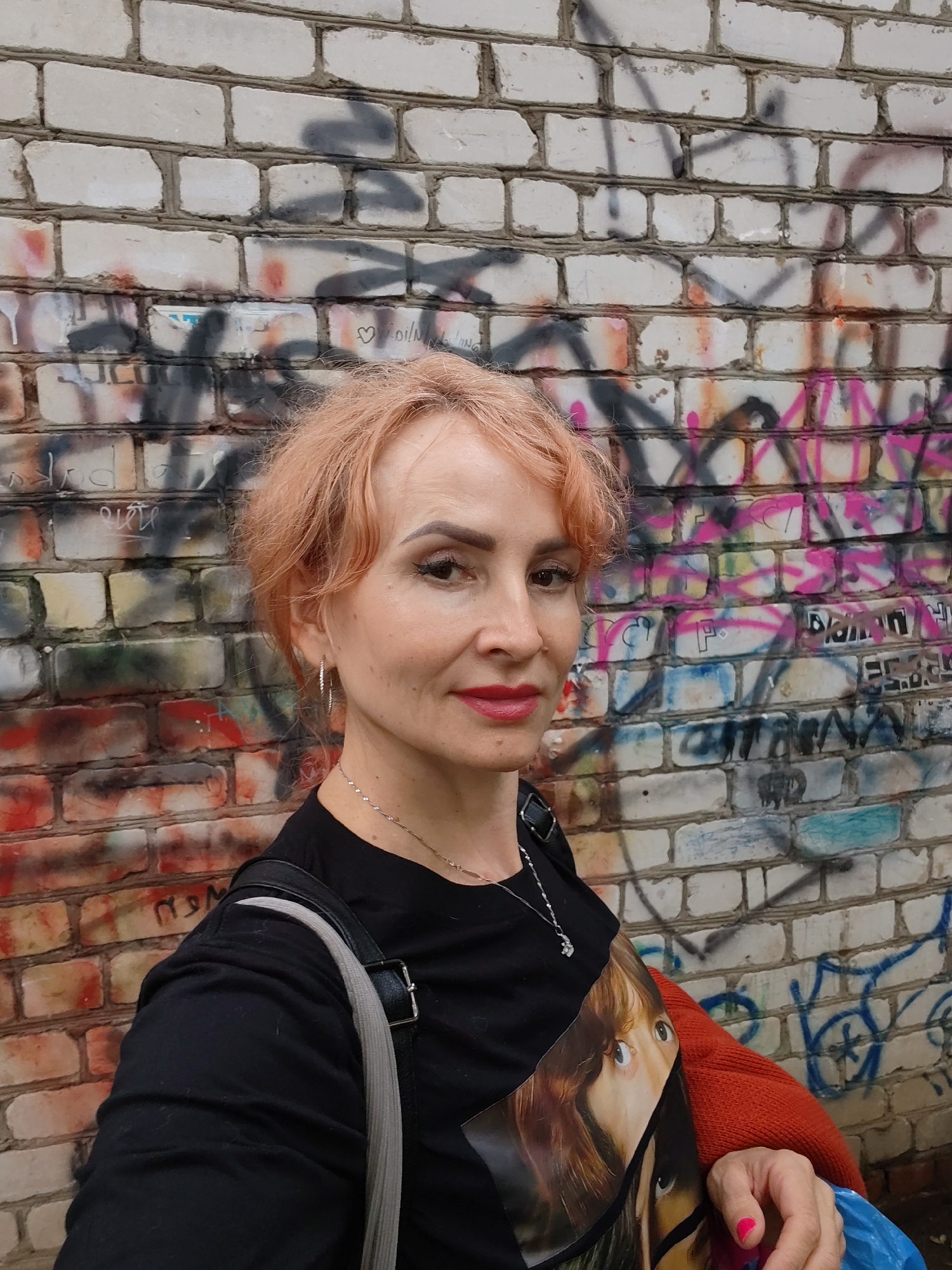 A middle-aged Caucasian woman with short, light pink hair stands in front of a graffiti-covered brick wall. She is wearing a black t-shirt with a graphic print of eyes and a small silver necklace. She has on hoop earrings and red lipstick. A grey strap from a bag is visible over her shoulder, and she is wearing an orange cardigan. Her left arm is bent, with her hand resting on her chest. A bright pink fingernail is visible. The wall behind her is made of white bricks, with various graffiti tags and drawings in multiple colors, including black, pink, red, yellow, and blue. The environment appears to be outdoors, possibly in an urban setting in Chaikovsky, Russia.