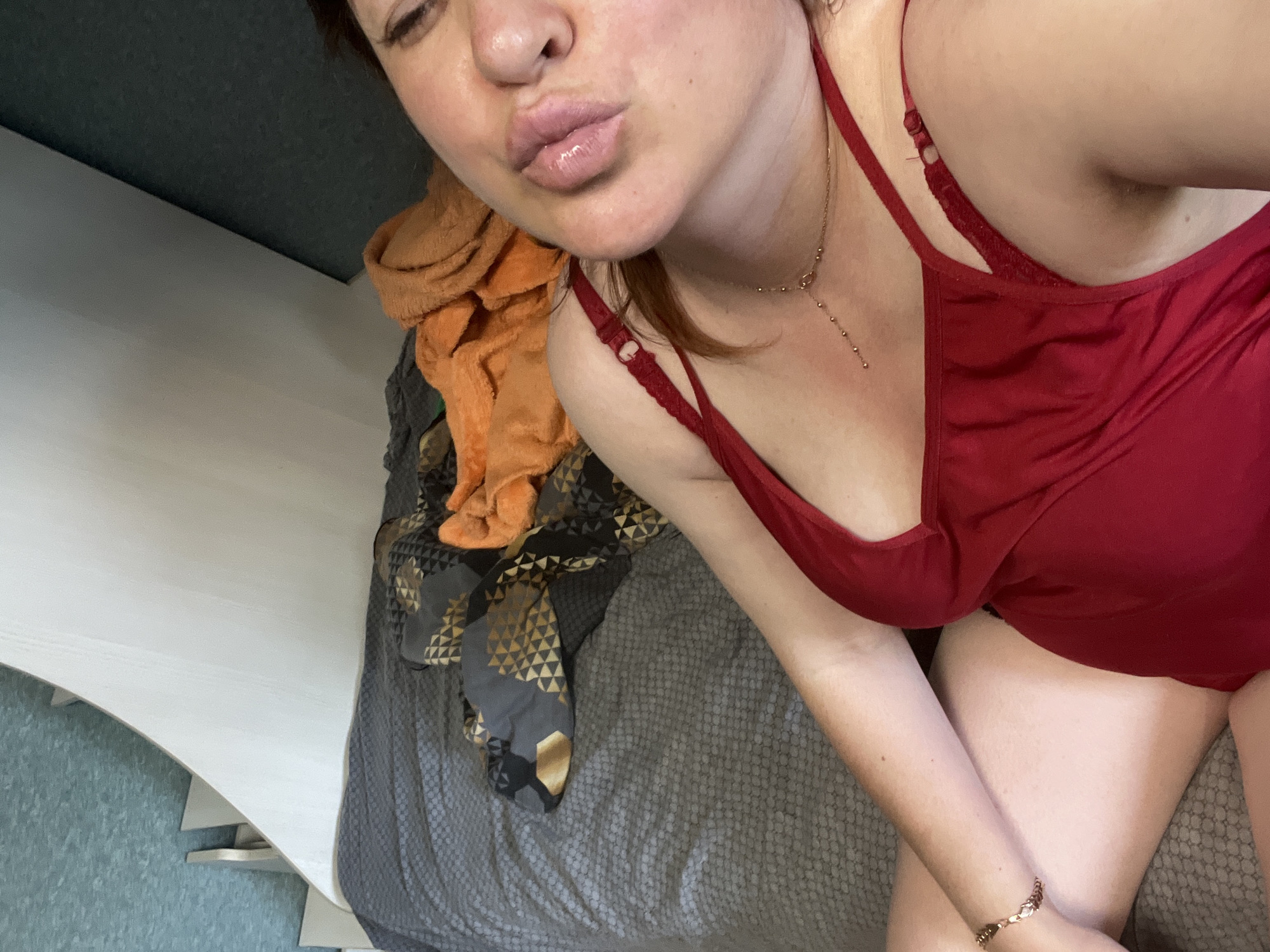 The image shows a close-up, high-angle, selfie-style shot of a woman lying on a bed. She is wearing a red top and a delicate gold necklace. Her eyes are closed and her lips are pursed, suggesting she is blowing a kiss. The bed is covered with a grey textured blanket, and there are piles of folded clothes, including orange and patterned fabrics, on the bed. The background features a textured wall with a geometric pattern and a white angled structure. The image does not provide any information about the specific location in the city of Noviy, Russia, or any details that would suggest an urban environment.