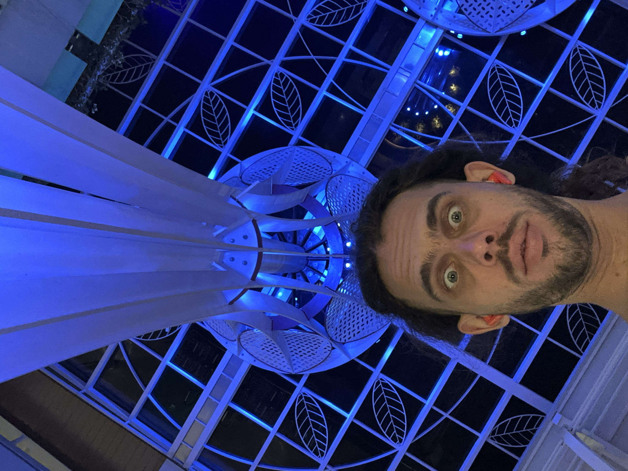 The image is a selfie taken from a low angle by a man with curly brown hair and a beard, looking directly at the camera with wide, startled eyes. He has bright orange earplugs in both ears. The background is dominated by a blue-lit architectural structure, possibly a ceiling or a large atrium, with a geometric pattern of white lines and leaf motifs. The man's face fills the bottom half of the frame, and the architectural elements are seen from above. The blue lighting suggests it is nighttime or an indoor space with artificial lighting. The location is identified as Đà Nẵng, Vietnam.