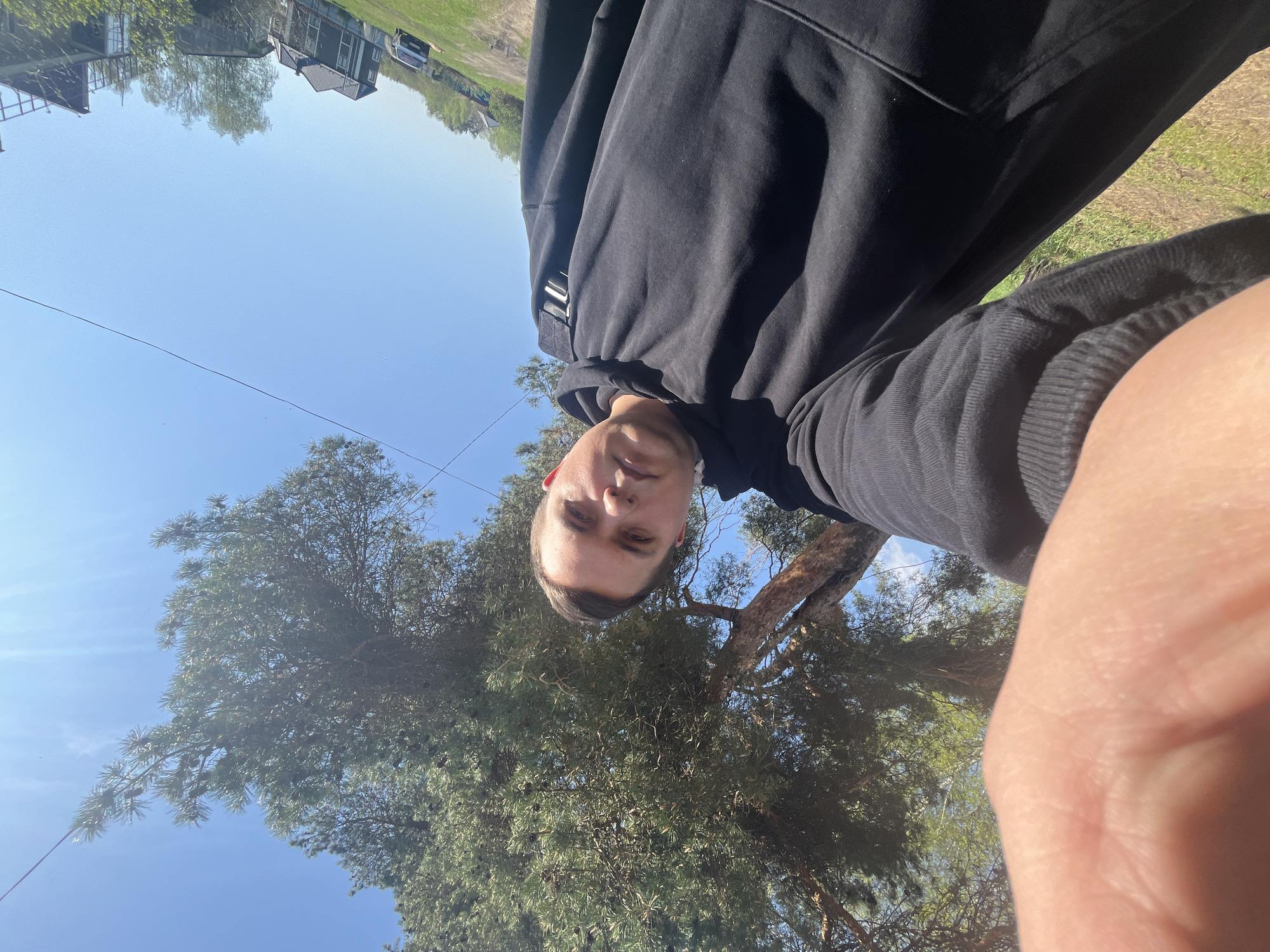 A low-angle selfie of a fair-skinned person with short, light-brown hair is positioned against a background of trees and a clear blue sky. The person is wearing a black hooded sweatshirt. A finger is prominently visible in the lower right corner of the frame. Behind the subject and trees, at the top of the image, some rooftops and a car can be seen. The scene suggests an outdoor setting. The city of Раздолье, Russia, is not overtly identifiable in this image.