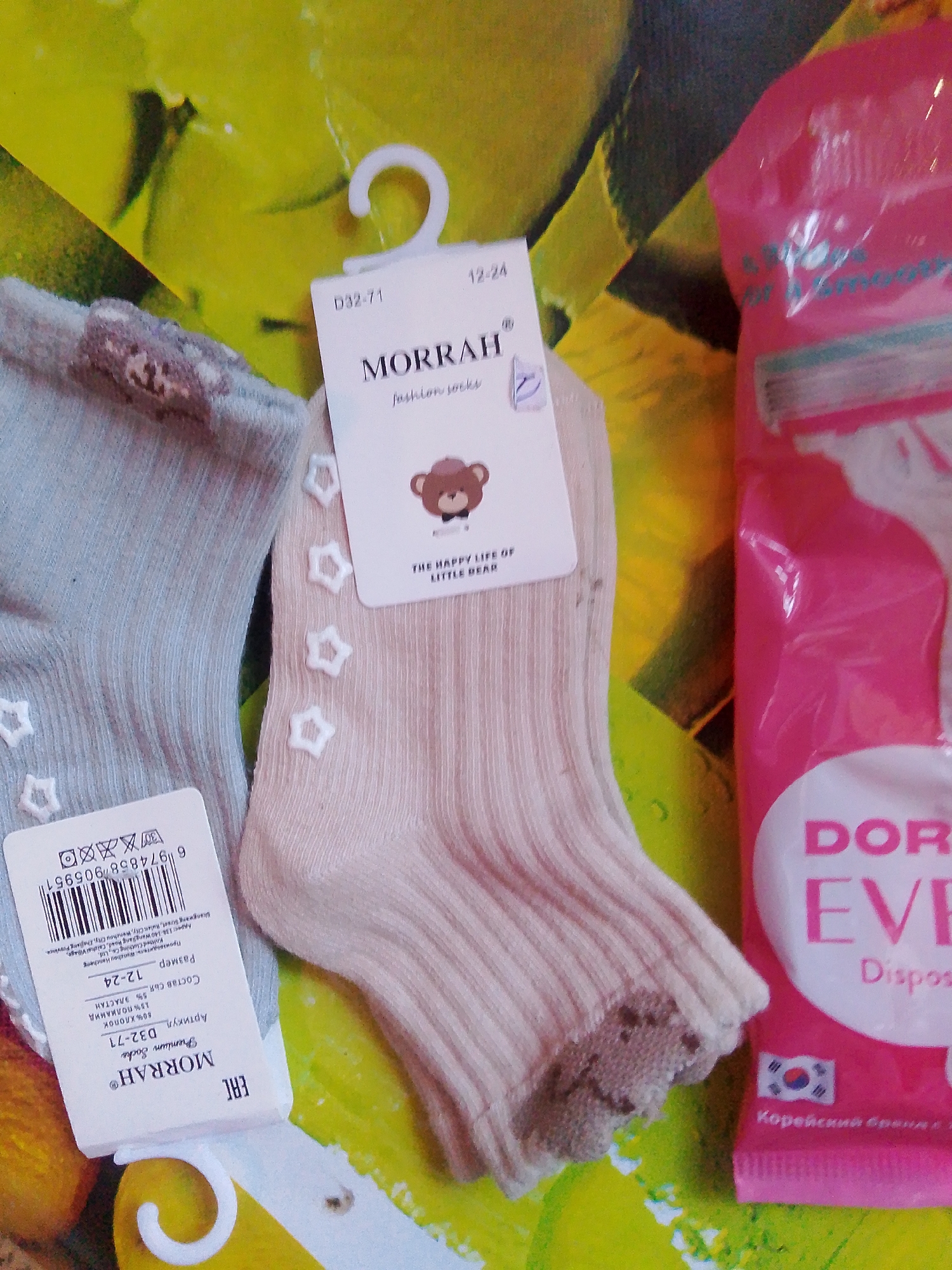 In a top-down shot, two pairs of baby socks are displayed on a surface with a yellow and green abstract pattern. The socks are light grey and light pink, both with a small embroidered teddy bear and small white stars. One sock is partially obscured by a white tag with "MORRAH" printed on it, along with product information. To the right of the socks, a bright pink package of what appears to be disposable cosmetic products is visible. The background suggests an indoor setting, though no specific city information like Хрестівка, Ukraine, can be deduced from the image.