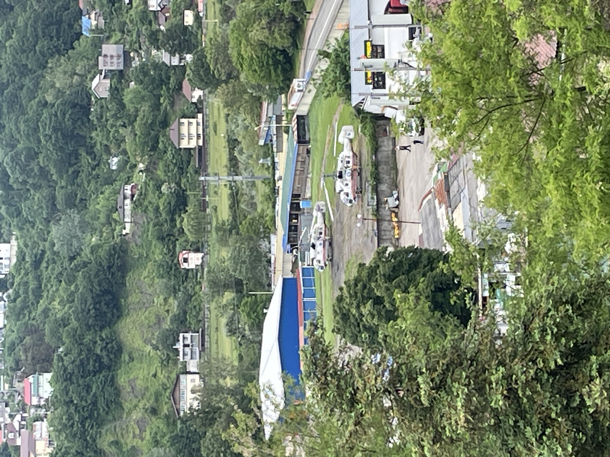 The image presents an elevated view of a scene likely located in the city of Sochi, Russia, based on contextual analysis. The landscape is a blend of natural and urban elements.
The left side of the frame is dominated by a densely vegetated hillside dotted with residential buildings, typical of a mountainous terrain. Moving towards the center, a cluster of low-rise buildings with various rooftop structures are visible. These appear to be commercial or industrial in nature, possibly related to helicopter maintenance.
Two helicopters are stationed on a tarmac or helipad. Adjacent to the helipad, a rectangular structure, possibly a hangar, is partially visible. A service road or highway runs parallel to the building complex.
On the right, lush greenery obscures a portion of the scene, suggesting the viewpoint is from a higher elevation, perhaps a hillside or building. Two human figures are visible standing between the commercial buildings and the helicopters.