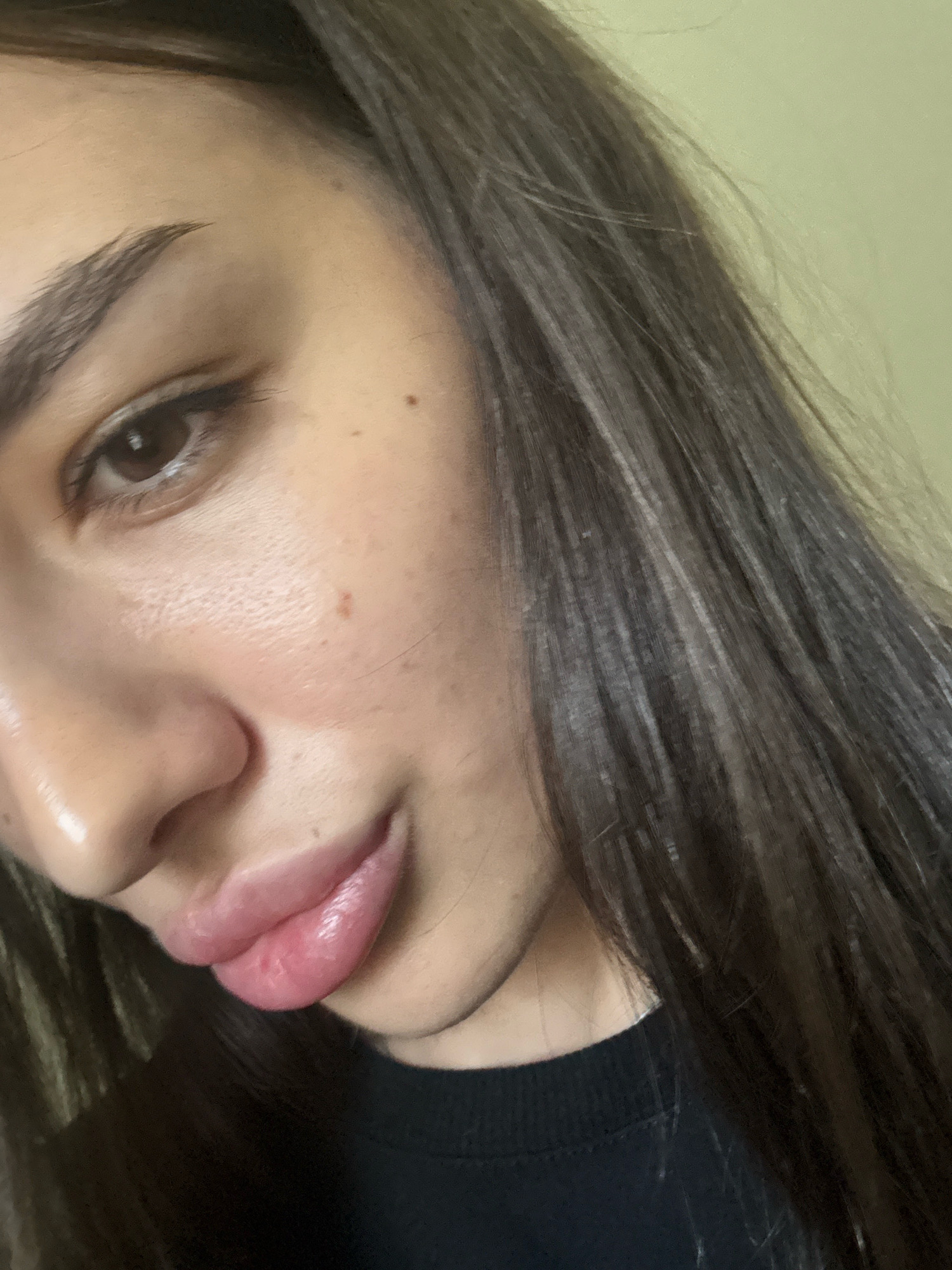 The image is a close-up portrait of a young woman's face, cropped to focus on her left eye, nose, and lips. She has dark hair that is long and slightly wavy, and it frames the right side of her face. Her skin appears clear with a few small moles, and she is wearing subtle makeup, with eyeliner visible on her eye and a glossy lip product. She is wearing a black t-shirt. The background is a plain, light-colored wall. There is no indication of a specific location within Chișinău, Moldova, or any other city. The image is a studio-style portrait.