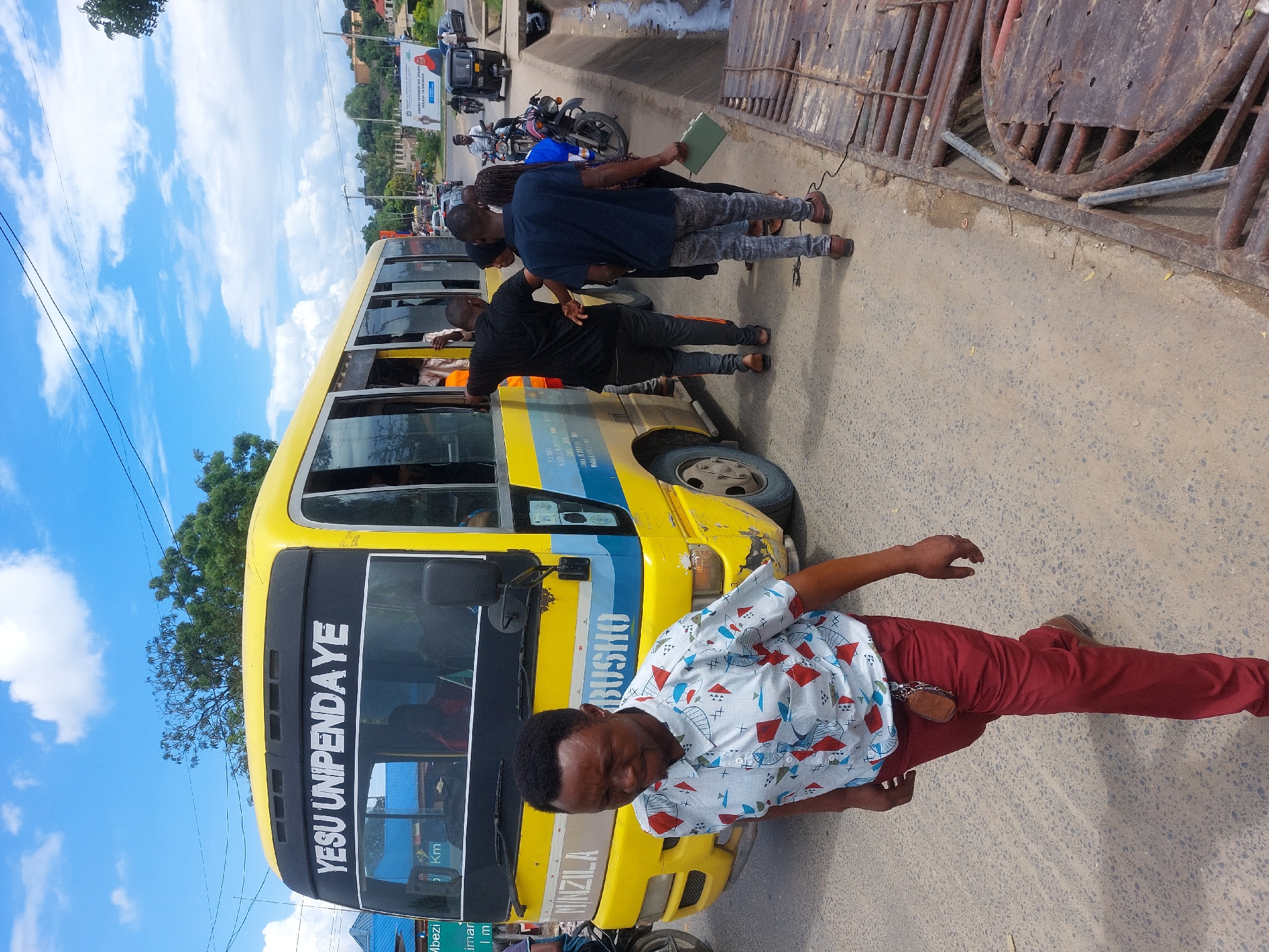 The image shows a yellow and blue minibus in Dar es-Salaam, Tanzania. "YESU UNIPENDA YE" is printed on the front of the minibus. Several people are standing around the bus, including a man in a patterned shirt and red pants. A motorcyclist is visible behind the bus. An elevated metal structure is on the right side of the image. The sky is partly cloudy.