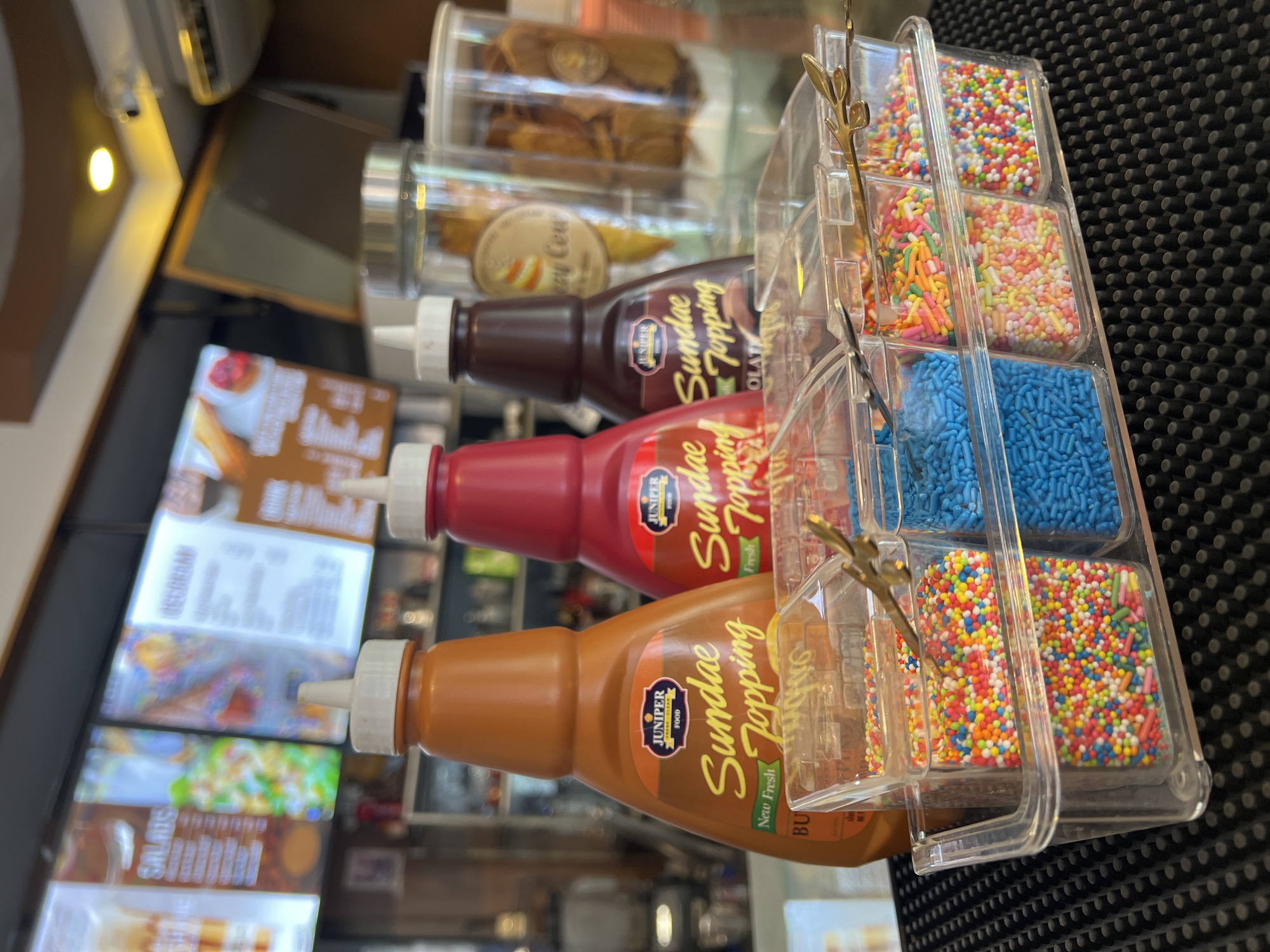 On a countertop, possibly in Baan Bo Phut, Thailand, sits an array of sundae toppings. There are four squeeze bottles of Juniper Food brand sundae toppings, flavors include butterscotch, strawberry, and chocolate. Two clear containers hold dehydrated mango and other crispy treats, with a sticker on the bottom-most container reading "Crazy Cow". Adjacent to these are four clear plastic bins holding sprinkles of different colors and sizes, including round rainbow sprinkles, pastel rod-shaped sprinkles, and dark blue rod-shaped sprinkles. A black mat is visible in the background. Food menus are seen in the background.