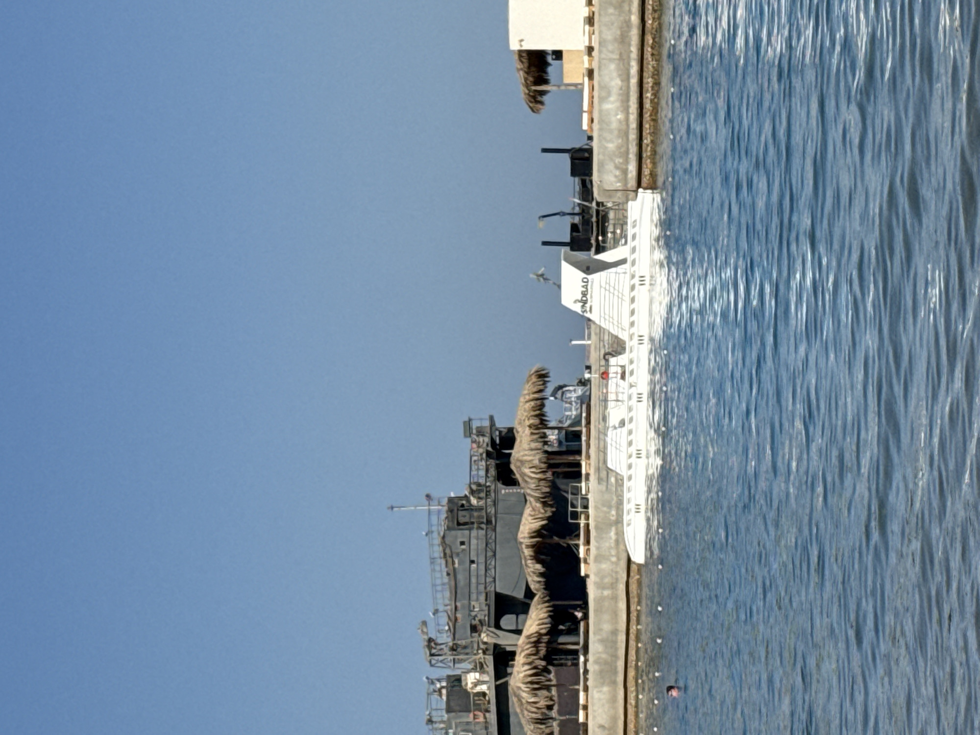 The image captures a daytime scene in Hurghada, Egypt, with a clear blue sky suggesting fair weather. The focus is on several boats docked along a concrete pier. In the foreground, a white boat, identified by the visible text "SINDBAD," is prominent. It appears to be a passenger vessel, possibly a ferry or tour boat, with multiple decks and windows. Further down the pier, a darker, larger vessel is docked, its structure more industrial in appearance, with scaffolding and what seem to be covered sections, possibly for cargo or protection. The water is a vibrant blue, showing ripples and reflections from the sun, indicating a calm sea. The overall setting is a harbor or marina, a typical coastal scene in a resort town like Hurghada. The presence of boats suggests maritime activity, likely related to tourism or transport. There are no people clearly visible in the image, adding to a sense of quietness at this moment.