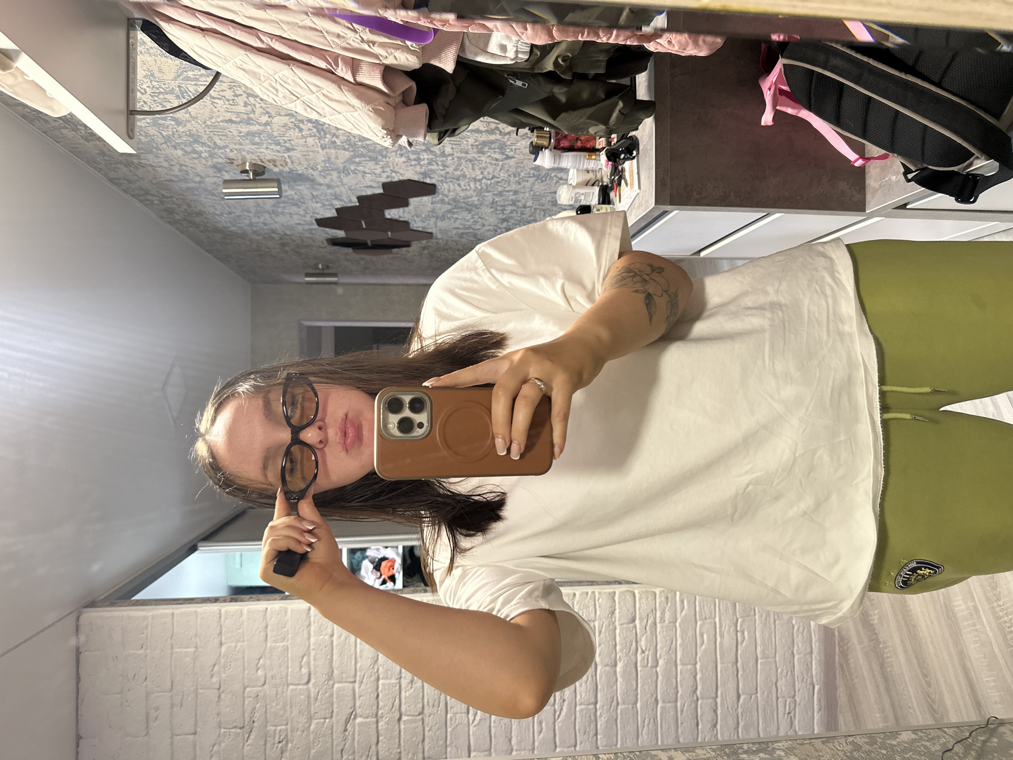 A woman is standing indoors, facing a mirror, holding a phone in her right hand and a pair of eyeglasses in her left hand. She is wearing glasses, a white t-shirt, and green sweatpants. Her right arm has a tattoo. The background includes a tiled wall, clothing items hanging in a closet, and a patterned ceiling. The city of Chaikovsky, Russia is not discernible from the provided visual information.