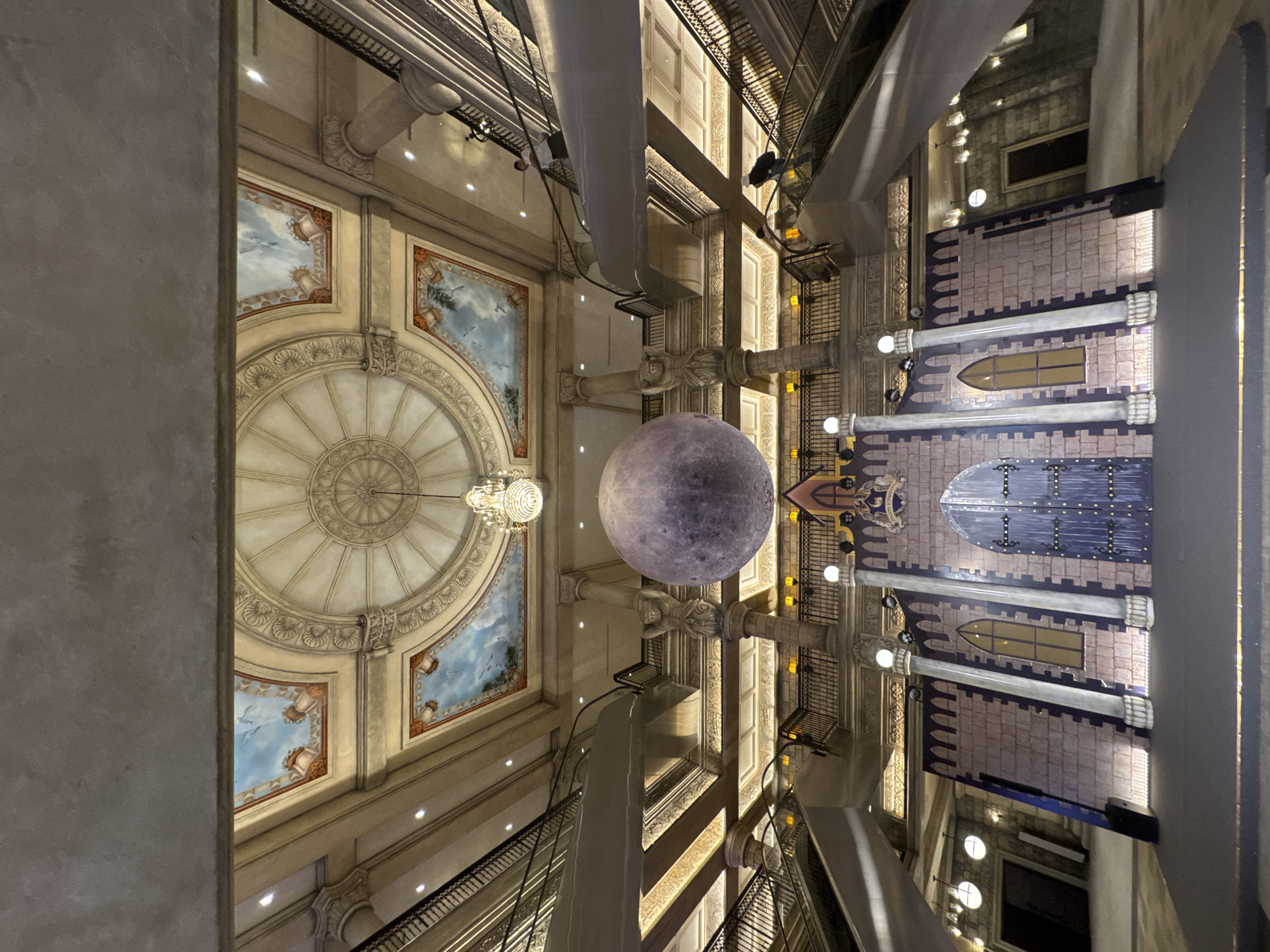 The image displays a grand, multi-story indoor space, likely an atrium or hall, captured from a lower level looking upwards. A prominent spherical object, resembling a moon with detailed craters, hangs centrally within the vast area. To its upper left, an ornate crystal chandelier is suspended from a richly decorated domed ceiling. This ceiling features intricate classical patterns and painted panels depicting blue skies with clouds and classical urns.
On the right side of the space, a large facade is designed to mimic a medieval or fantasy castle. It showcases textured stone-like walls, arched windows, and imposing dark blue double doors adorned with metal studs. A crest with an indistinguishable emblem is visible above the doors.
The architecture throughout is elaborate, characterized by multiple levels of balconies or walkways supported by ornate columns with carved capitals, and walls featuring detailed classical moldings and paneling. Modern spotlights are integrated to illuminate the scene, complementing the artificial lighting that pervades the indoor setting. No individuals are visible. The overall aesthetic combines classical grandeur with whimsical, fantastical elements, creating an impression of a lavish commercial, hotel, or entertainment complex in Da Nang, Vietnam. No discernible text is clearly visible.