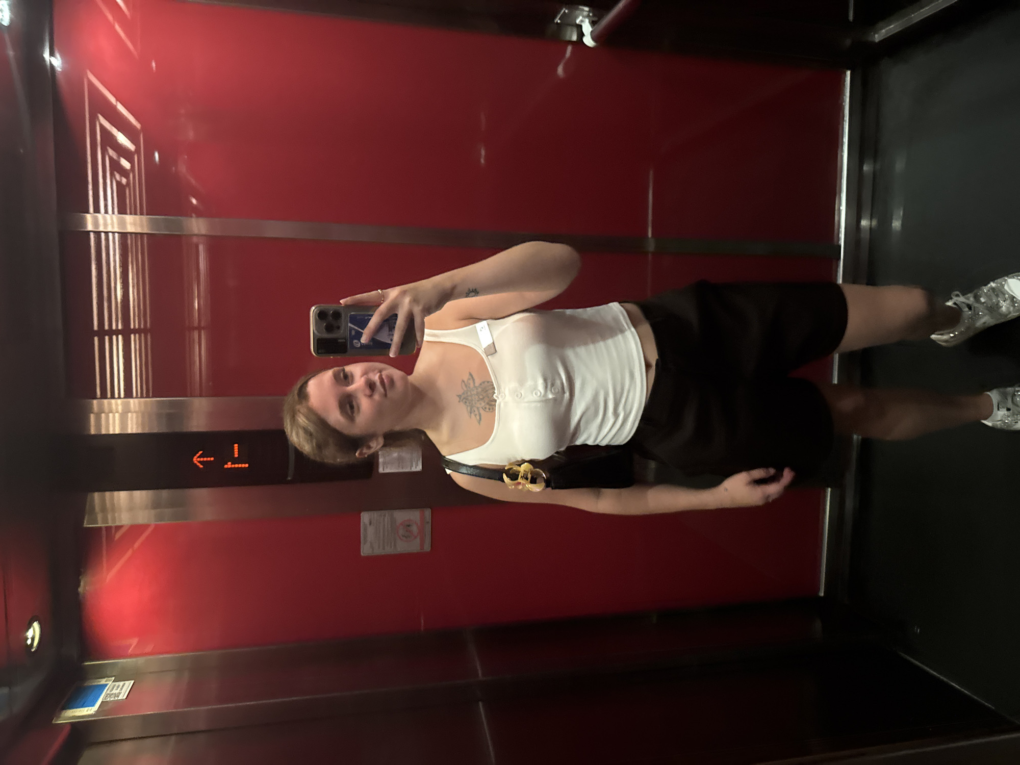 A young woman is standing in an elevator, taking a selfie. The elevator has red walls and silver trim. The woman is wearing a white tank top, black shorts, and white sneakers. She has a black purse slung over her shoulder and is holding her phone in her right hand. There is a tattoo on her chest and another on her left arm. The elevator floor is black. There is a sign on the elevator wall with an upward-pointing arrow. The image is taken from a slightly low angle.