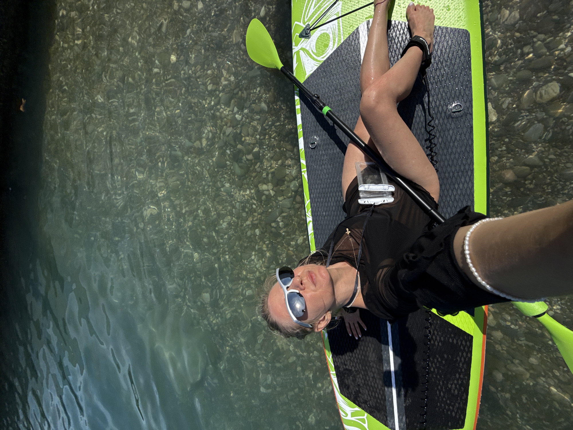 A woman is lying on a paddleboard in shallow, clear water with a rocky bottom. She is wearing sunglasses, a black sheer top, and black shorts. A paddle is lying on the board next to her, and a leash is attached to her ankle and the board. The water reflects the sunlight, and the rocks are visible through the surface. The scene appears to be a recreational activity on a sunny day.