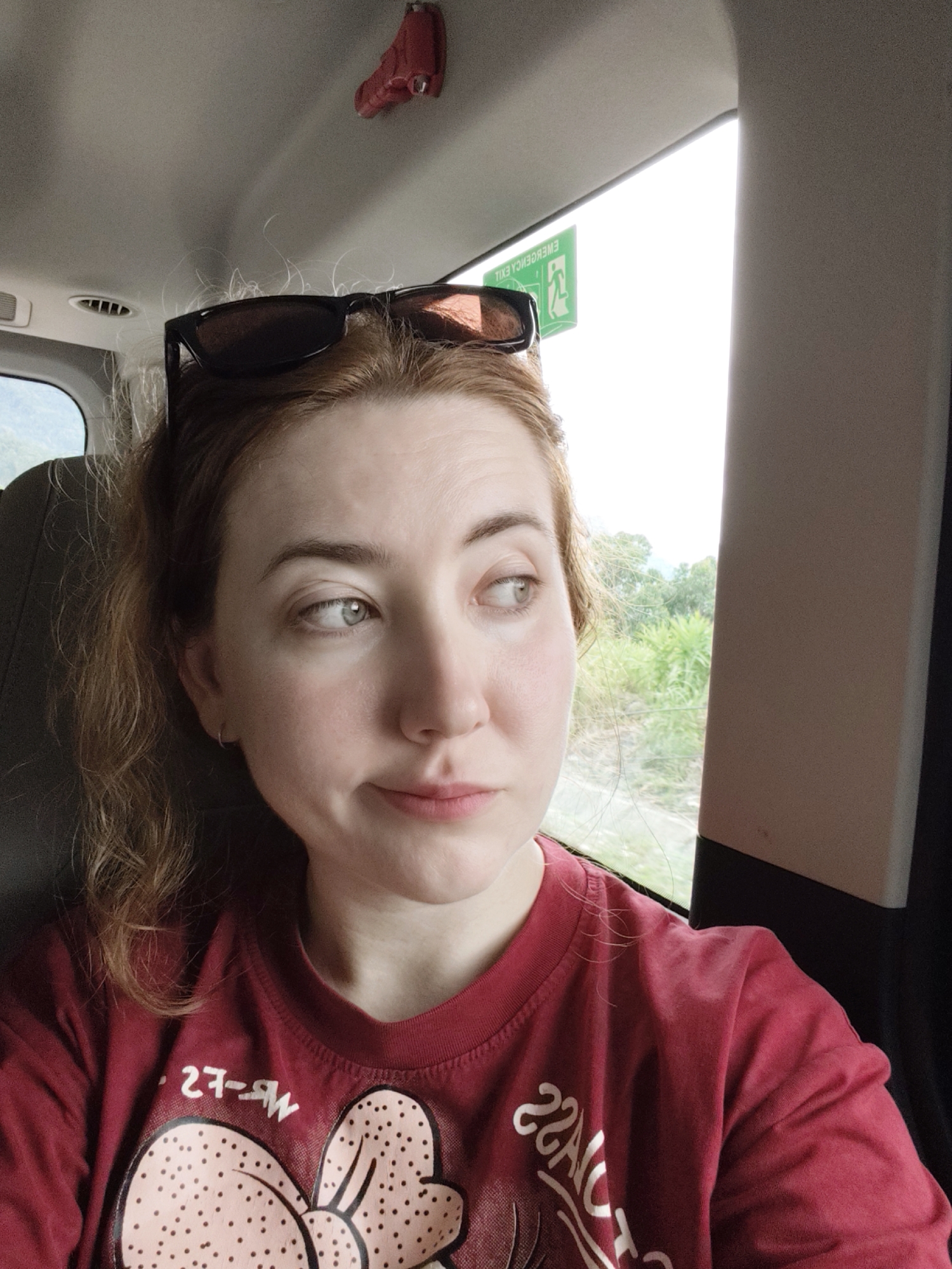 A young woman with light skin and auburn hair sits in the passenger seat of a vehicle, looking out the window to her right. She is wearing a red t-shirt with a graphic design featuring a large bow with polka dots. Sunglasses are perched on top of her head. A small red object is attached to the ceiling of the vehicle above her. A green emergency exit sign is visible on the window. The background outside the window shows lush green foliage and a bright sky. The image is taken from a medium shot, focusing on the woman's face and upper body.