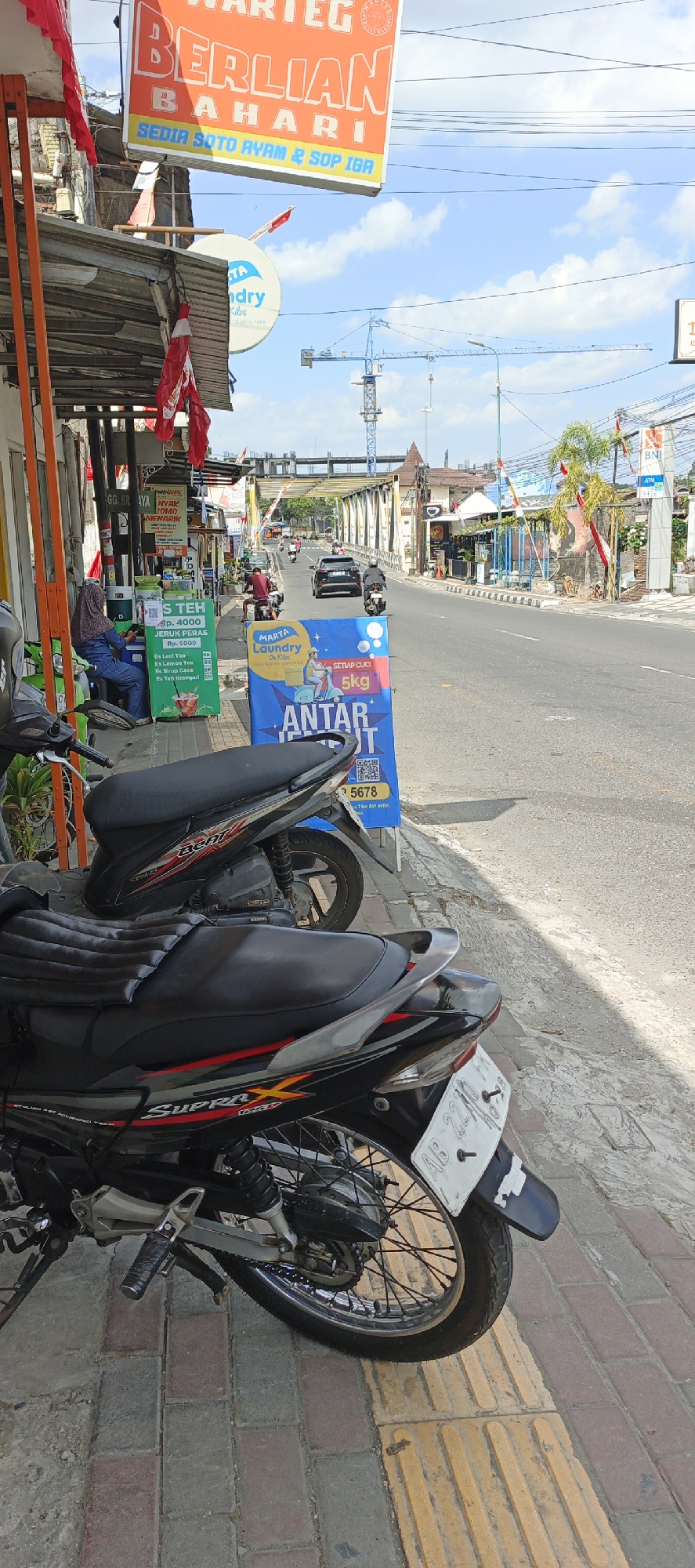 A street scene in Yogyakarta, Indonesia, features two parked motorcycles on a sidewalk beside a road. A large orange sign overhead reads "WARTEG BERLIAN BAHARI SEDIA SOTO AYAM & SOP IGA." A blue sign for "MARTA Laundry" is on the sidewalk, advertising "5 KG ANTAR JEMPUT." In the background, traffic moves along the road, with cars and motorcycles. A construction crane is visible in the distance. The sky is partly cloudy. A person sits on a green stall on the sidewalk.