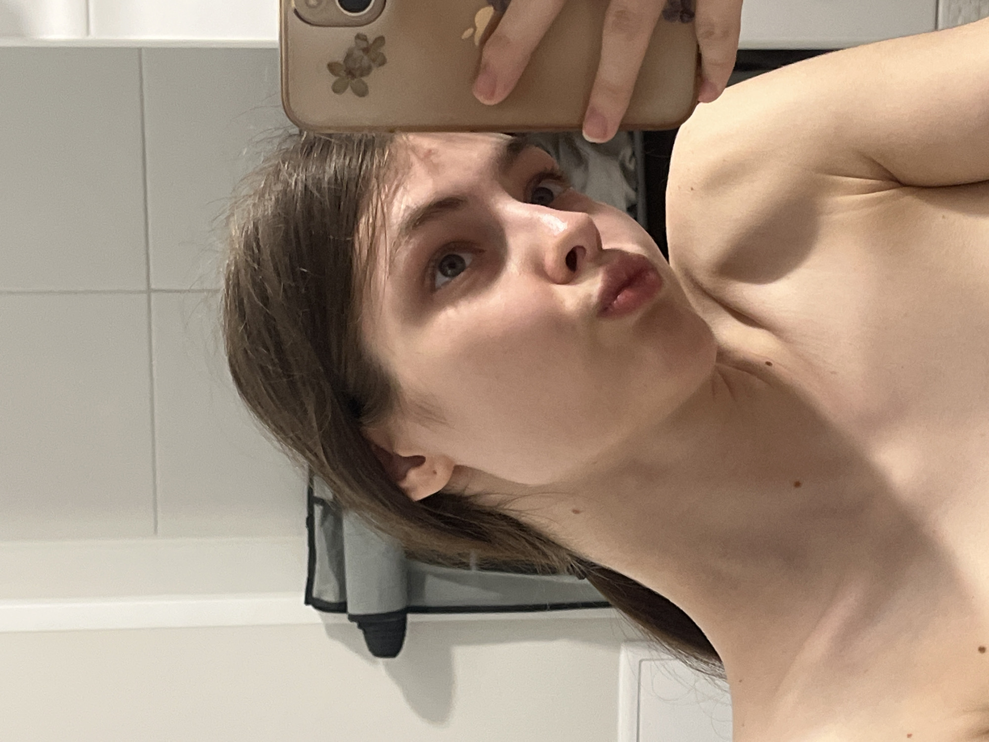 The image depicts a young adult female taking a selfie in what appears to be a mirror, positioned in an indoor environment in the city of Saint Petersburg, Russia. The subject, with fair skin and brown hair pulled back, is shown from the upper chest upwards. Her head is tilted to the right, and her lips are pursed in a "pout" expression, with her left eye gazing upwards. She holds a light-colored smartphone, featuring a decorative case with a floral pattern and an Apple logo, which obscures the right side of her face. Her bare shoulders and neck are visible, displaying several small, scattered moles. A partial view of a tattoo is visible on a finger of her right hand, which is holding the phone. The background consists of plain, light-colored tiled walls, with a grey and black hanging storage item visible behind her left shoulder.