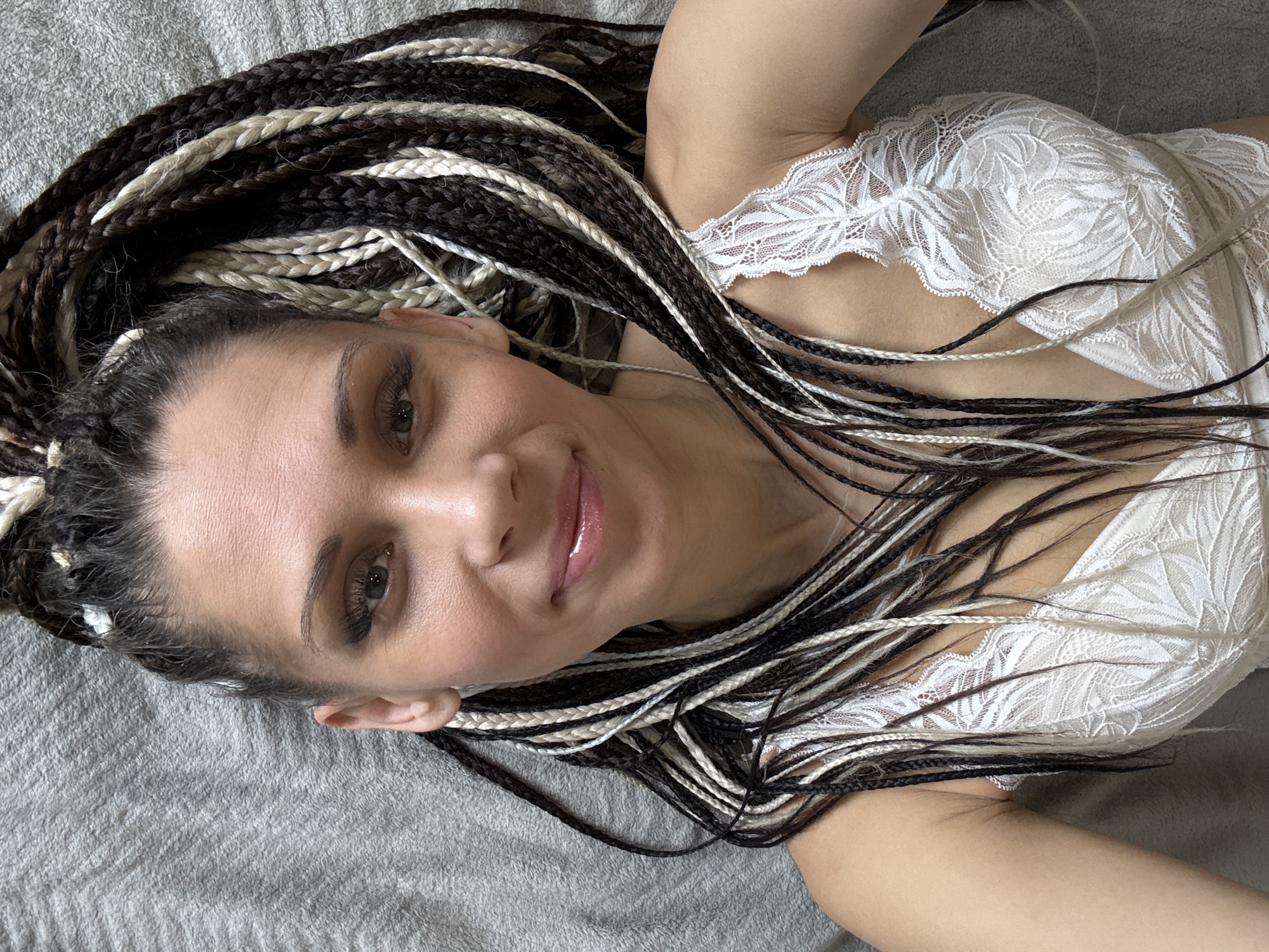 A woman with braids is lying on a gray surface. The braids are black, white, and brown. She is wearing a white lace top. She is smiling slightly. The photo was taken from above. The city of Fedorino, Russia is not discernible in the image.