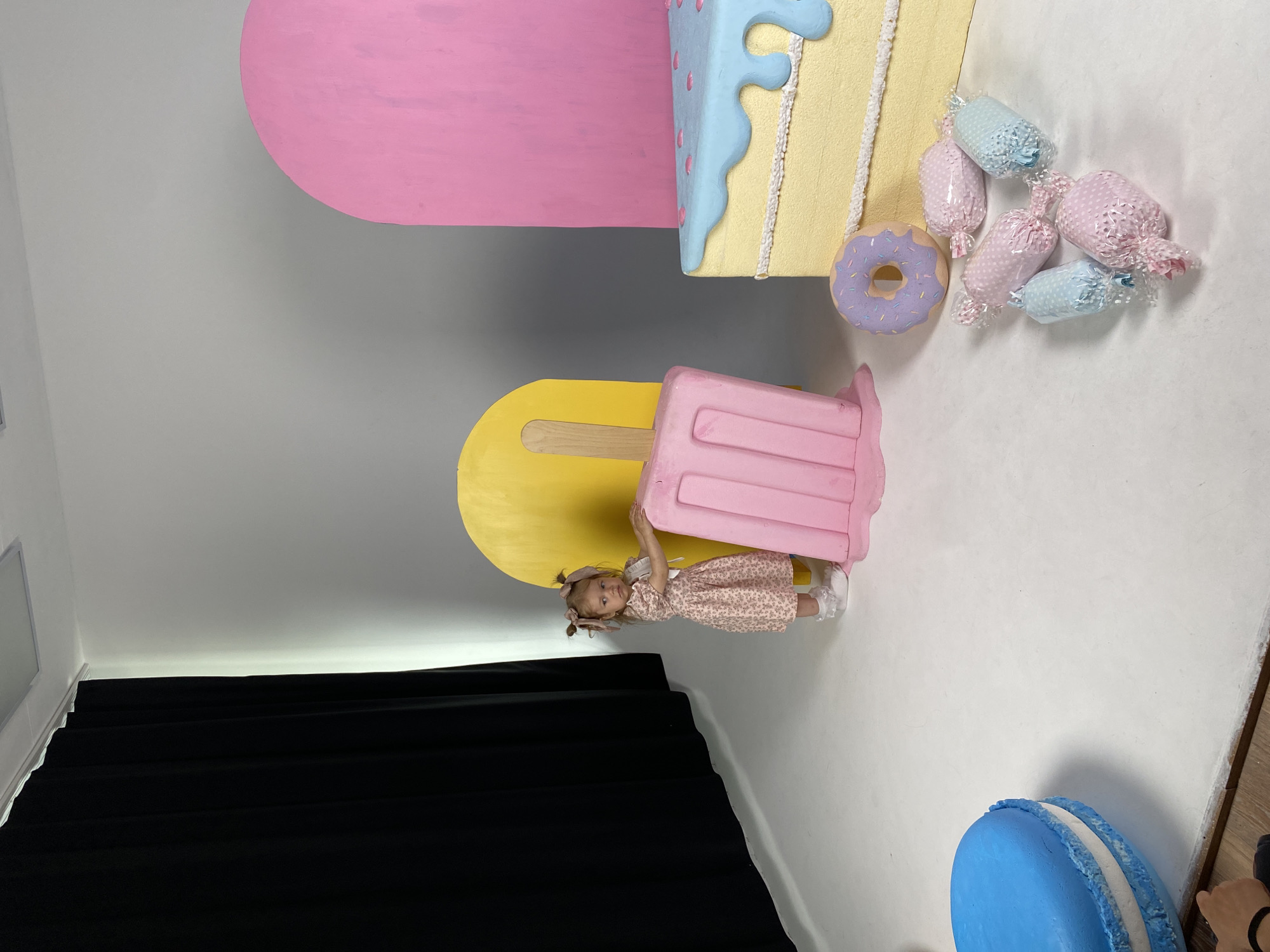 A young girl with blonde pigtails, wearing a pink floral dress and white socks, stands in a studio. She is leaning on a large pink popsicle prop. Behind her, a yellow arch shape is visible, and to her right, a pink arch shape and a large slice of cake prop are present. The cake prop is yellow with light blue frosting and pink sprinkles. In the foreground, a blue macaron prop is on the left. Several wrapped candies and a purple donut prop are scattered on the white floor. A black curtain is draped on the left side of the frame. The setting appears to be a photo studio with a white floor and gray backdrop. The city of Chaikovsky, Russia is not discernible in the image.