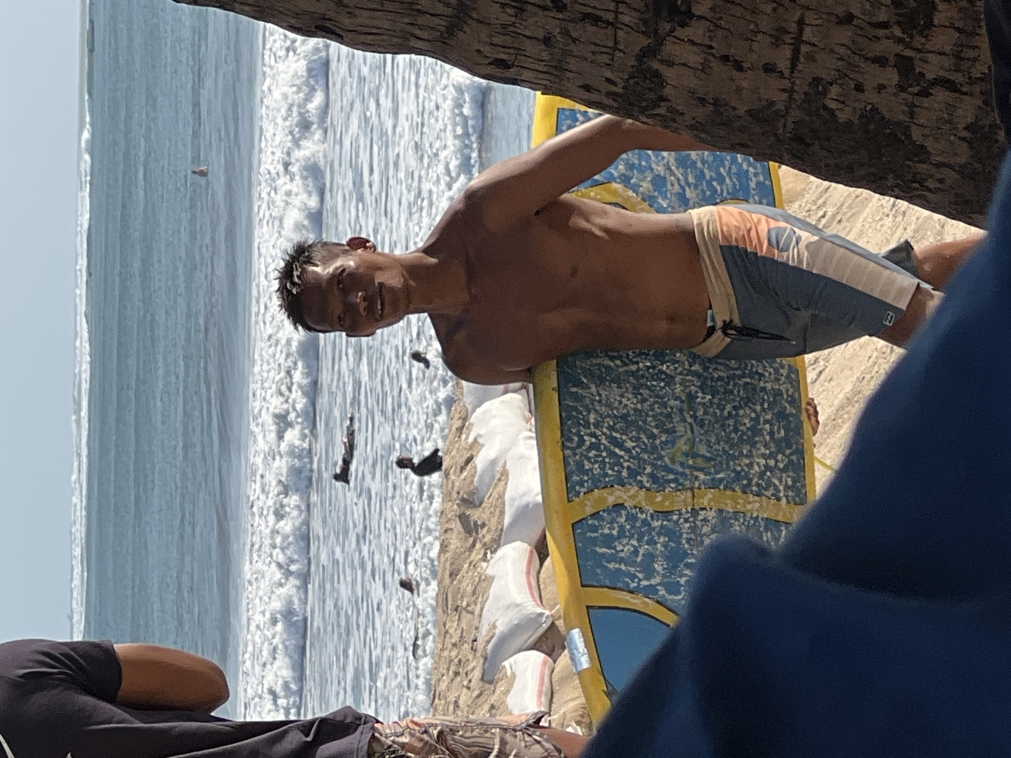 A shirtless man with short dark hair stands on a sandy beach in Kuta, Indonesia, holding a blue and yellow surfboard. He is wearing gray, white, and orange board shorts. The ocean with waves is visible behind him, and several other people are in the water. In the foreground, a blue object obstructs part of the view, and to the right, a tree trunk is partially visible. Sandbags are arranged on the beach behind the man and the surfboard. The sky is clear and bright, suggesting it is daytime.