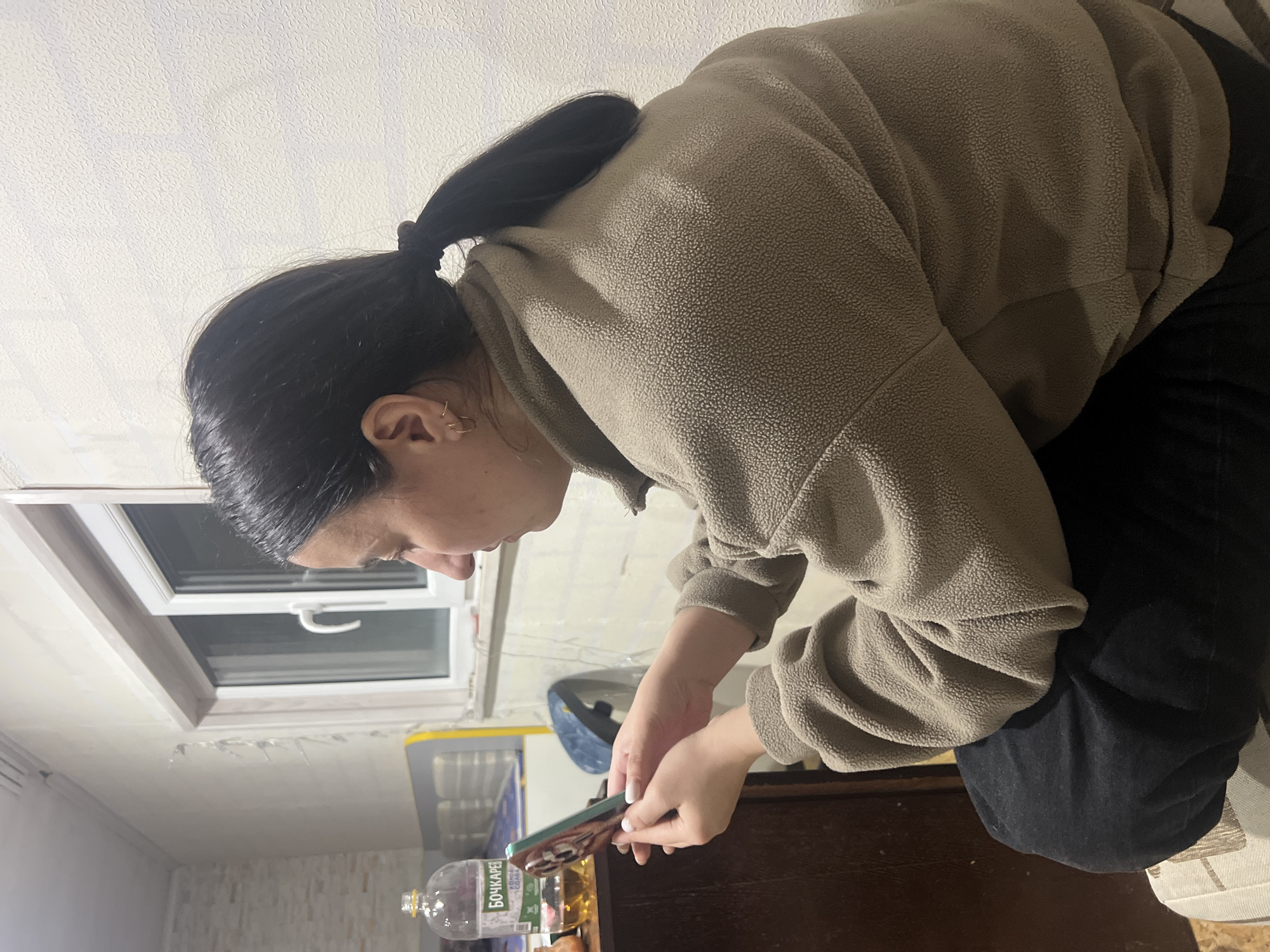 A woman with dark hair pulled back in a ponytail is looking at a smartphone in her hands. She is wearing a beige fleece jacket and black pants. The photo appears to be taken indoors, with a window visible in the background. A bottle labeled "БОЧКАРЕВ" can be seen near the bottom of the image, potentially indicating a location in Russia. Chaikovsky, Russia is the location.