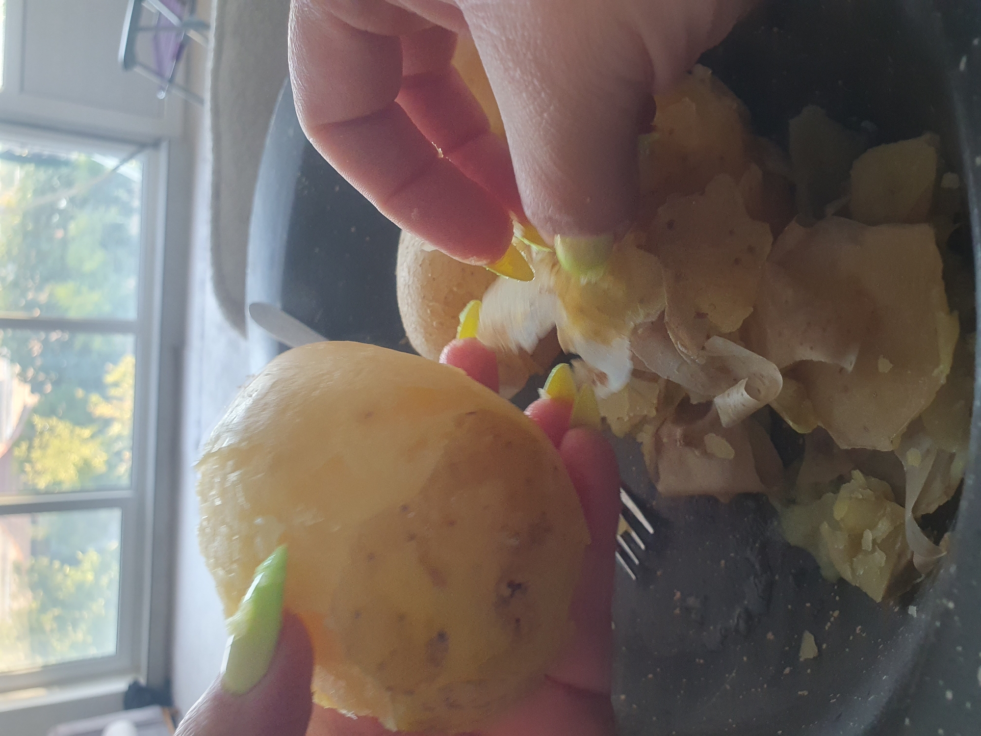 In a close-up shot, a person's hands with bright neon green painted nails are peeling a cooked potato over a dark bowl filled with peeled potatoes and their skins. The potato being held is round and pale yellow, with a few brown speckles. The other hand is actively peeling another potato, with pieces of skin curling away. The bowl appears to be made of speckled dark material and contains several chunks of cooked potato and loose skins. A fork with metal tines is partially visible in the lower left corner of the bowl. In the background, a large window reveals a blurry scene of green trees and buildings under a bright sky, suggesting an indoor setting with natural light. The overall impression is of food preparation. The location is not discernible from the image.