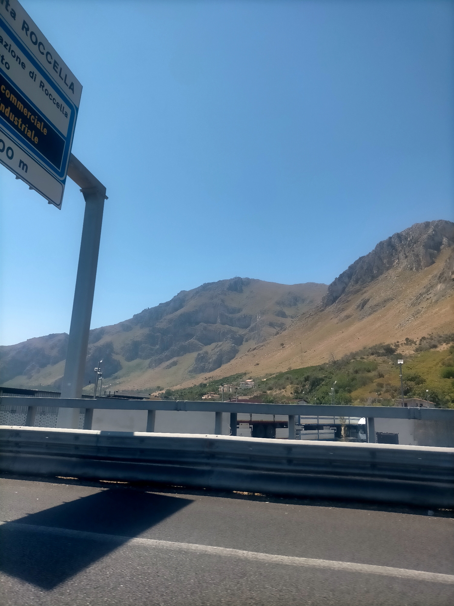 The image shows a view from a highway in Palermo, Italy, with a road barrier in the foreground and a mountain range in the background. A directional sign is visible on the left, indicating "ROCCELLA" and directions for "Commerciale Industriale" within 200 meters. The sky is clear blue, and the sun is shining, casting a shadow of the barrier onto the road. In the distance, there are some buildings nestled into the hillside. The scene appears to be a typical roadside view in a mountainous area.