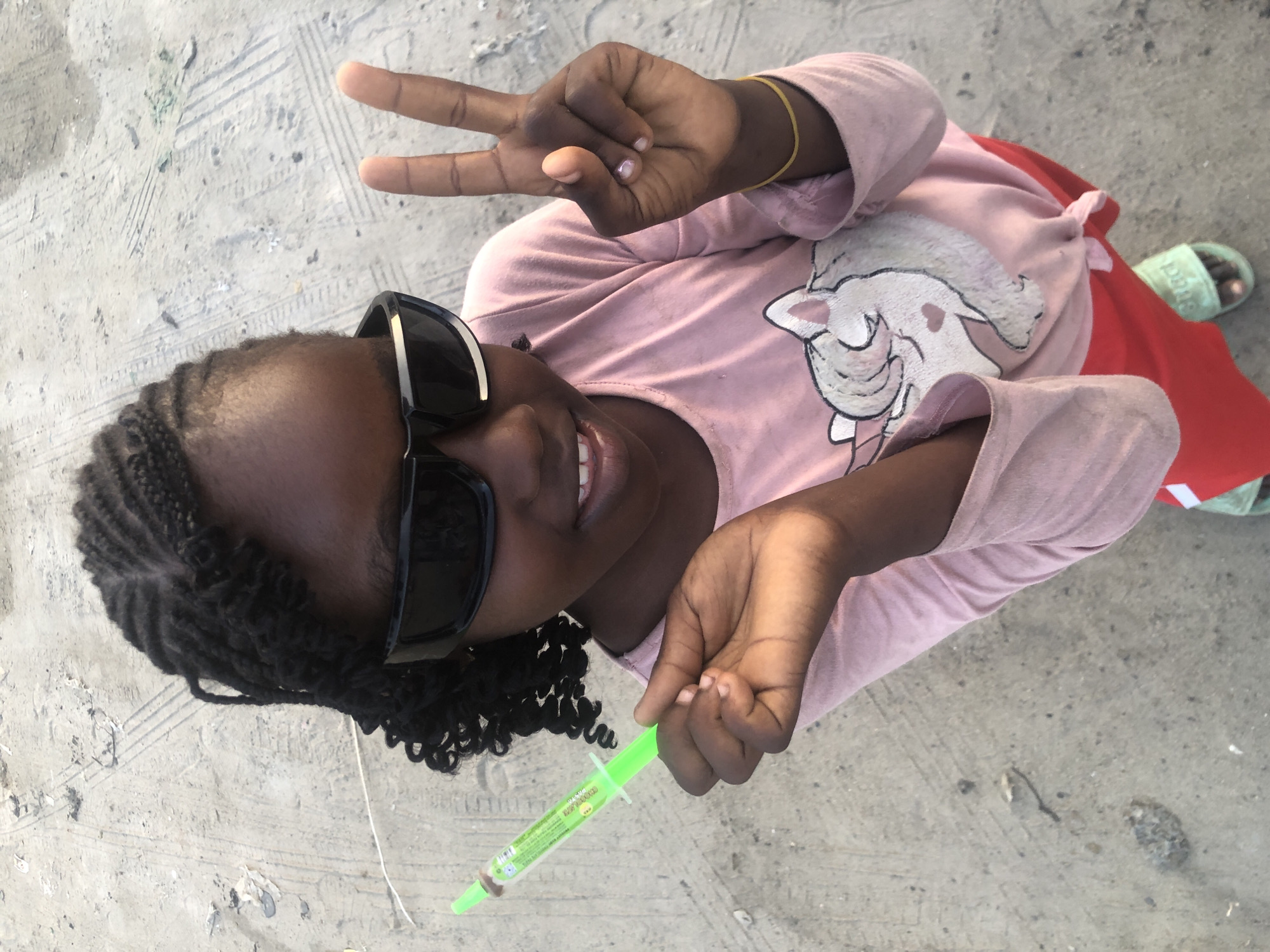 A young girl with dark skin, wearing black sunglasses and a pink long-sleeved shirt with a unicorn graphic, is holding a green syringe-shaped candy. She is making a peace sign with her right hand. The girl has braided hair, with some strands loose around her face and neck. She is wearing a red skirt and light green sandals. The background is a sandy or dirt ground with tire tracks visible. The scene appears to be outdoors in Ado, Nigeria.
