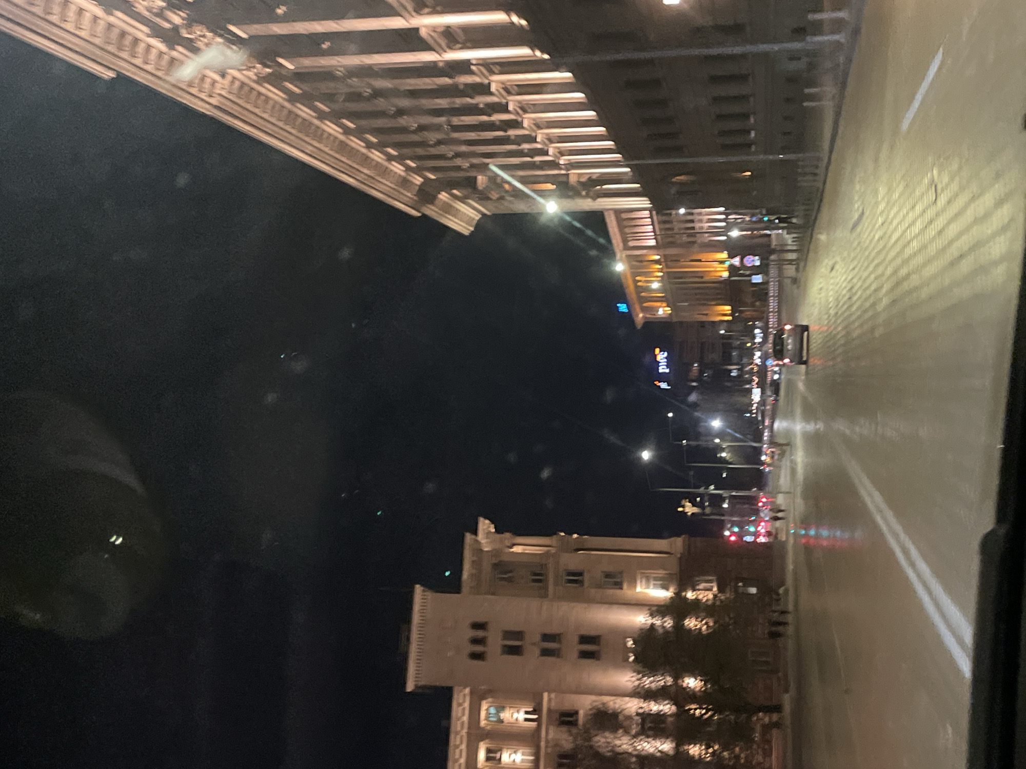 The image depicts a night scene on a street in Sofia, Bulgaria. Buildings are illuminated along the road, and traffic lights are visible in the distance. The street is lined with trees. The photo appears to be taken from inside a car. The architecture suggests a historical urban setting.
