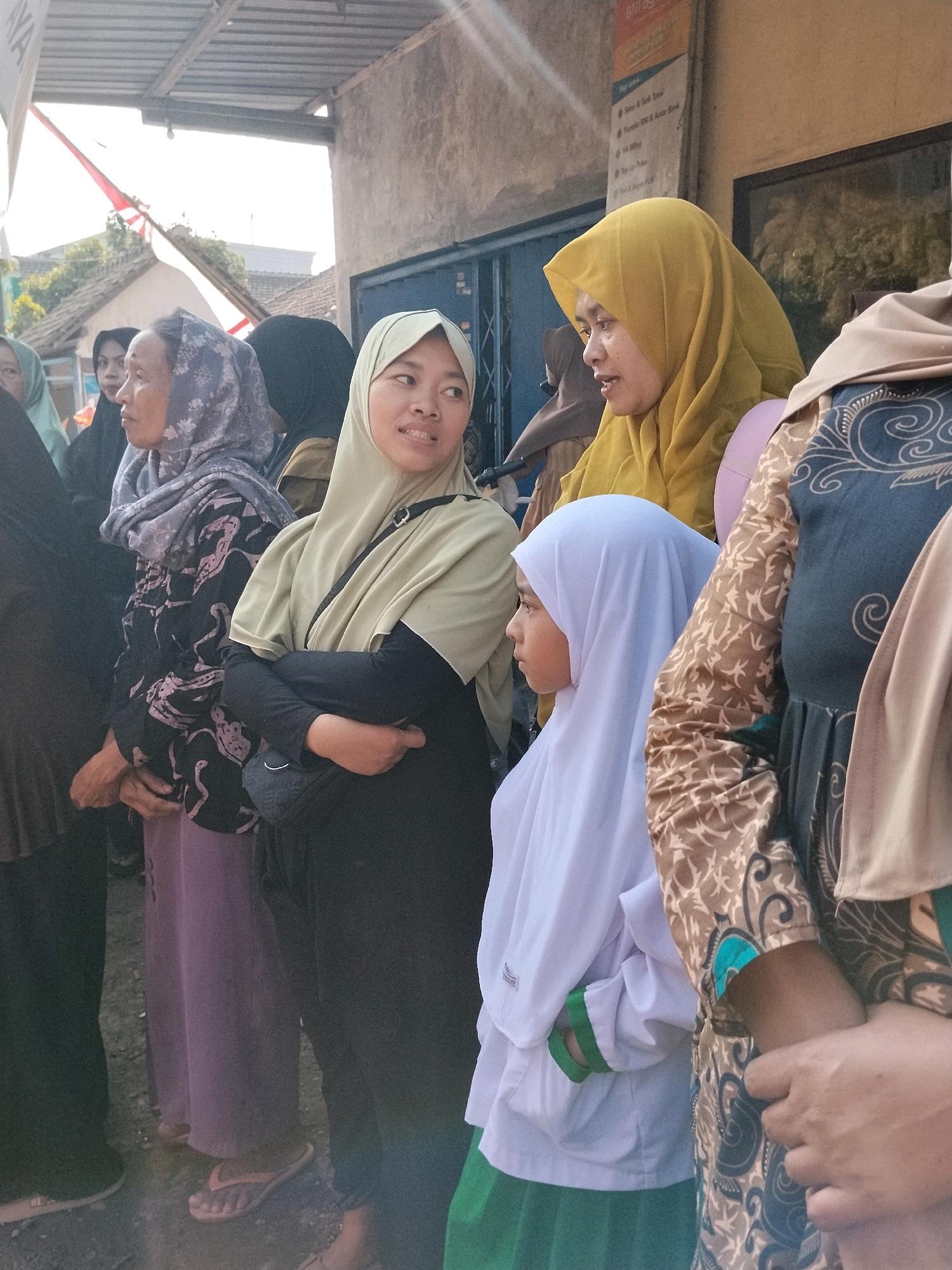 In Tlogosari, Indonesia, a group of women and a young girl, all wearing hijabs, are gathered outdoors. The woman in the foreground, wearing a light green hijab and a black outfit, has her arms crossed and is looking to her right. Next to her, a young girl in a white hijab and green skirt stands with her body turned to the left. Behind them, other women are partially visible, one in a yellow hijab and another in a patterned black and purple outfit. The environment appears to be near a building with a corrugated roof and a slightly textured wall. The lighting suggests it is daytime, with sunlight casting shadows.