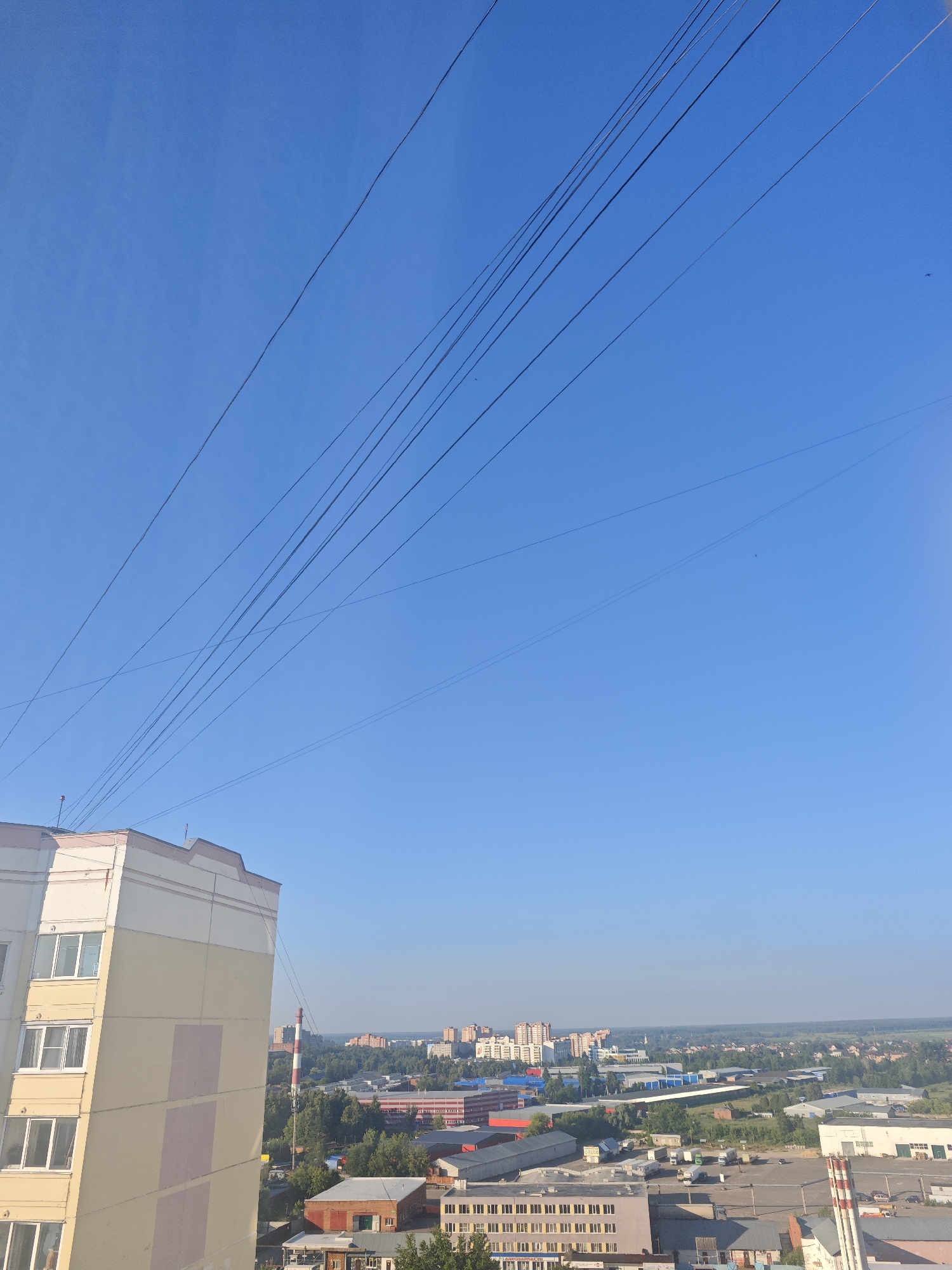 An aerial view captures the city of Voskresenski, Russia, on a clear day. The foreground features a portion of a tall, light-colored apartment building with beige and pale pink panels. Beyond the building, the cityscape unfolds with a variety of structures including industrial buildings with red roofs, smaller residential buildings, and a prominent tall chimney with red and white stripes. Dense green trees are interspersed throughout the urban landscape. Overhead, a series of thin, dark power lines crisscross the bright blue sky, creating a distinct visual element. The horizon is flat, suggesting a generally level terrain. The image appears to have been taken from a high vantage point, possibly another building.