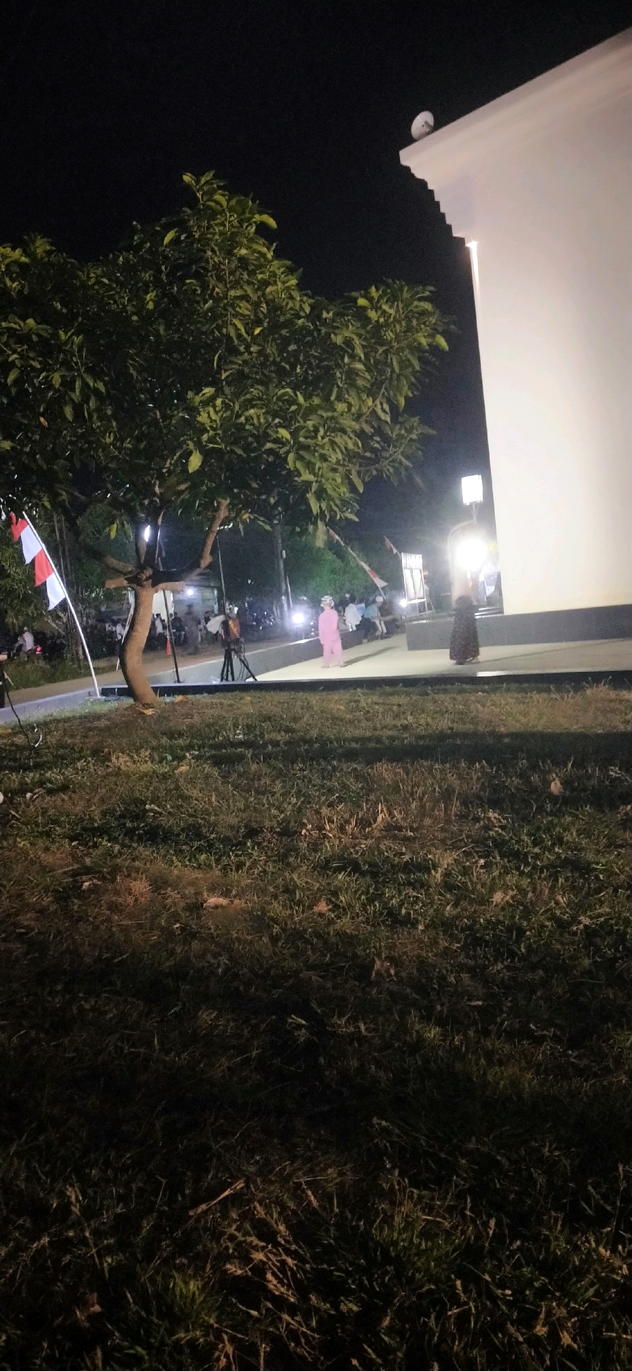 A group of people are gathered outdoors at night, near a white building with a satellite dish on its roof. A large tree with dark green leaves is visible in the foreground. Indonesian flags, red and white, are planted on poles to the left of the tree. A person in a pink outfit stands near the building, facing towards the left. Another person in a dark, patterned dress stands to the right of the pink-clad individual, facing towards the left. Several vehicles, including motorcycles, are parked in the background, with their headlights illuminating the scene. The ground in the foreground is covered in grass. The location appears to be an outdoor event or gathering.