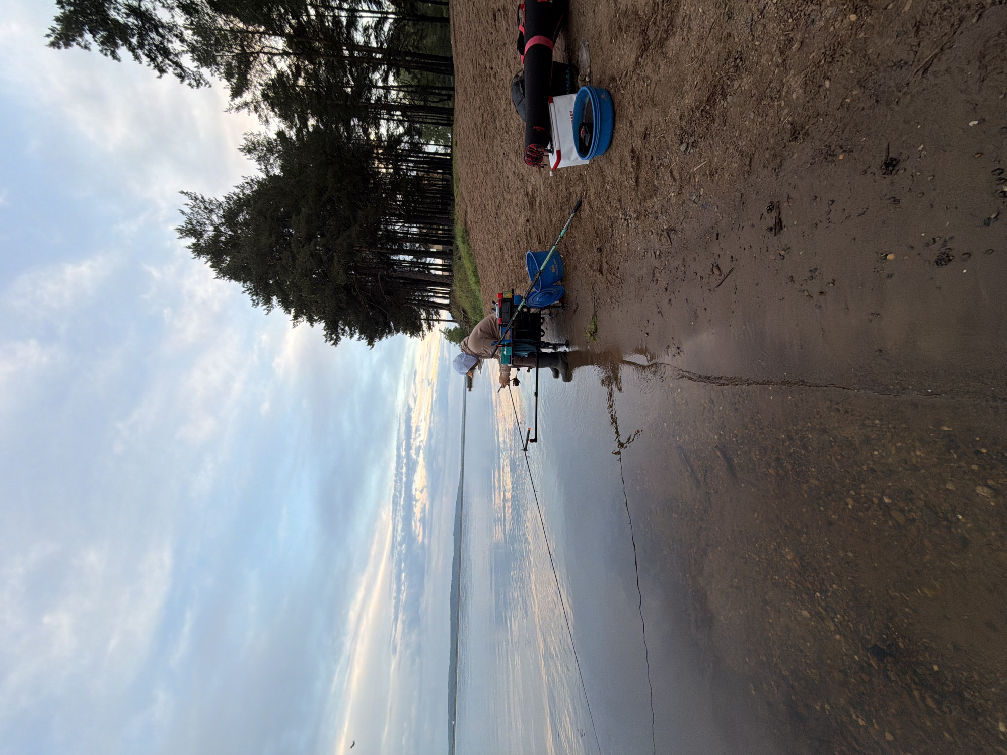An eye-level outdoor shot depicts a person fishing on the edge of a body of water, likely a lake or river, in what appears to be Chaikovsky, Russia. The person, clad in neutral-toned clothing and headwear, is bent over, attending to a fishing rod that extends into the water. Various fishing equipment, including buckets and bags, are arranged nearby on the shore. Behind the person, the shoreline is marked by a sandy area and a line of dense trees. The body of water takes up a significant portion of the frame, reflecting the sky. The sky exhibits a mix of clouds and clear patches.