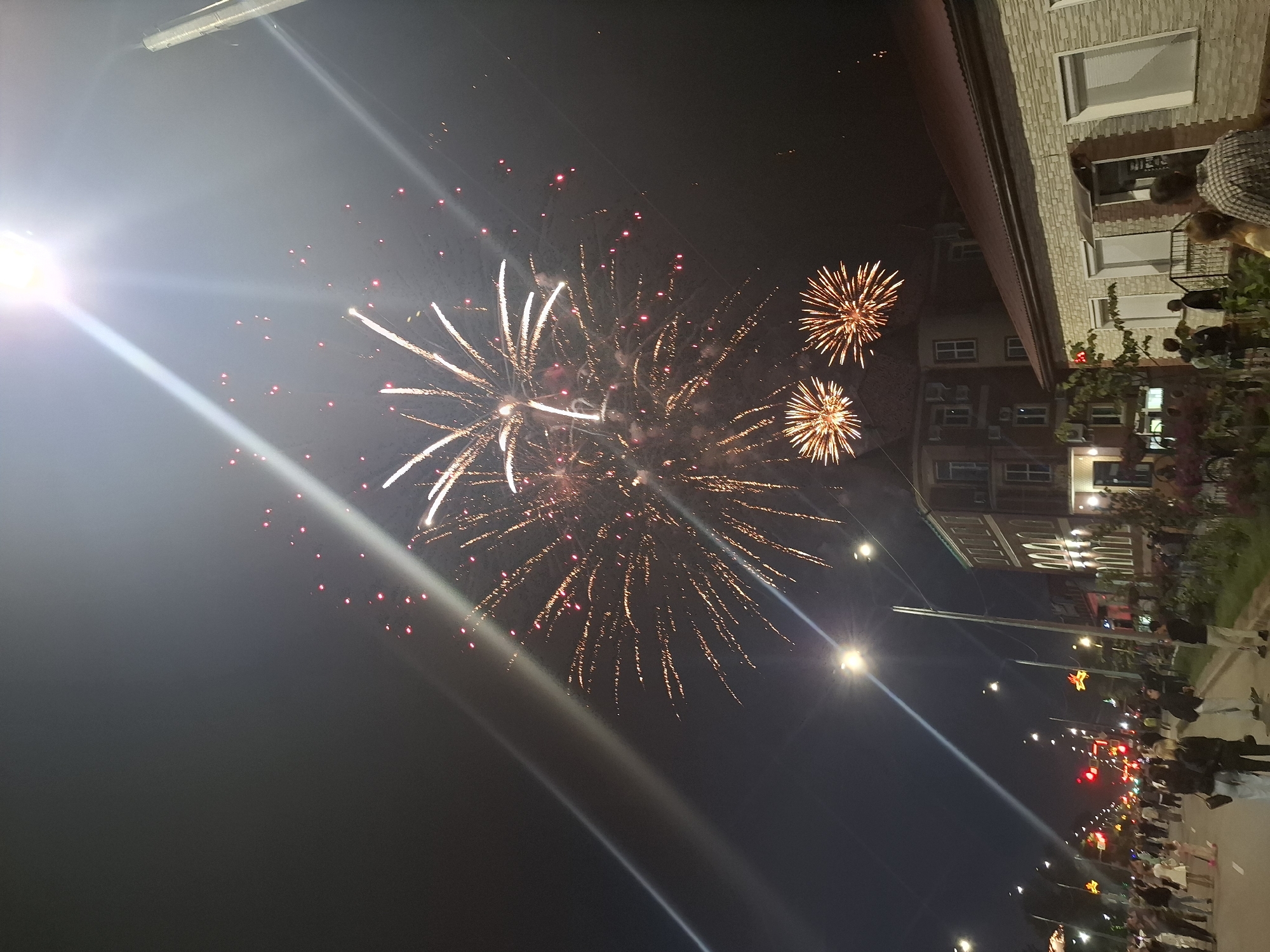 This image captures a nighttime scene in the city of Svobodny, Russia, featuring a fireworks display over a street lined with buildings and a crowd of onlookers. The sky is dark, illuminated by the bright bursts of fireworks. Prominent fireworks are visible in the upper portion of the image, with smaller explosions scattered throughout. A bright light source, likely a streetlamp or spotlight, creates strong lens flares and beams of light that extend diagonally across the sky.
The street is paved and appears to be an urban thoroughfare, with traffic lights and streetlights visible. Several buildings flank the street. On the right, a low-rise building with a light-colored stone facade and a reddish-brown roof is present. To its left, a taller brick building with multiple windows stands. On the left side of the street, more buildings and streetlights are visible, leading into the distance.
A crowd of people is gathered on the sidewalks and street, watching the fireworks. They are diverse in age and attire, with some individuals standing and others appearing to be walking. Some people are looking up at the sky.
The overall atmosphere is festive, suggesting a public celebration or event. The image is taken from a slightly elevated perspective, looking up towards the fireworks and down the street.