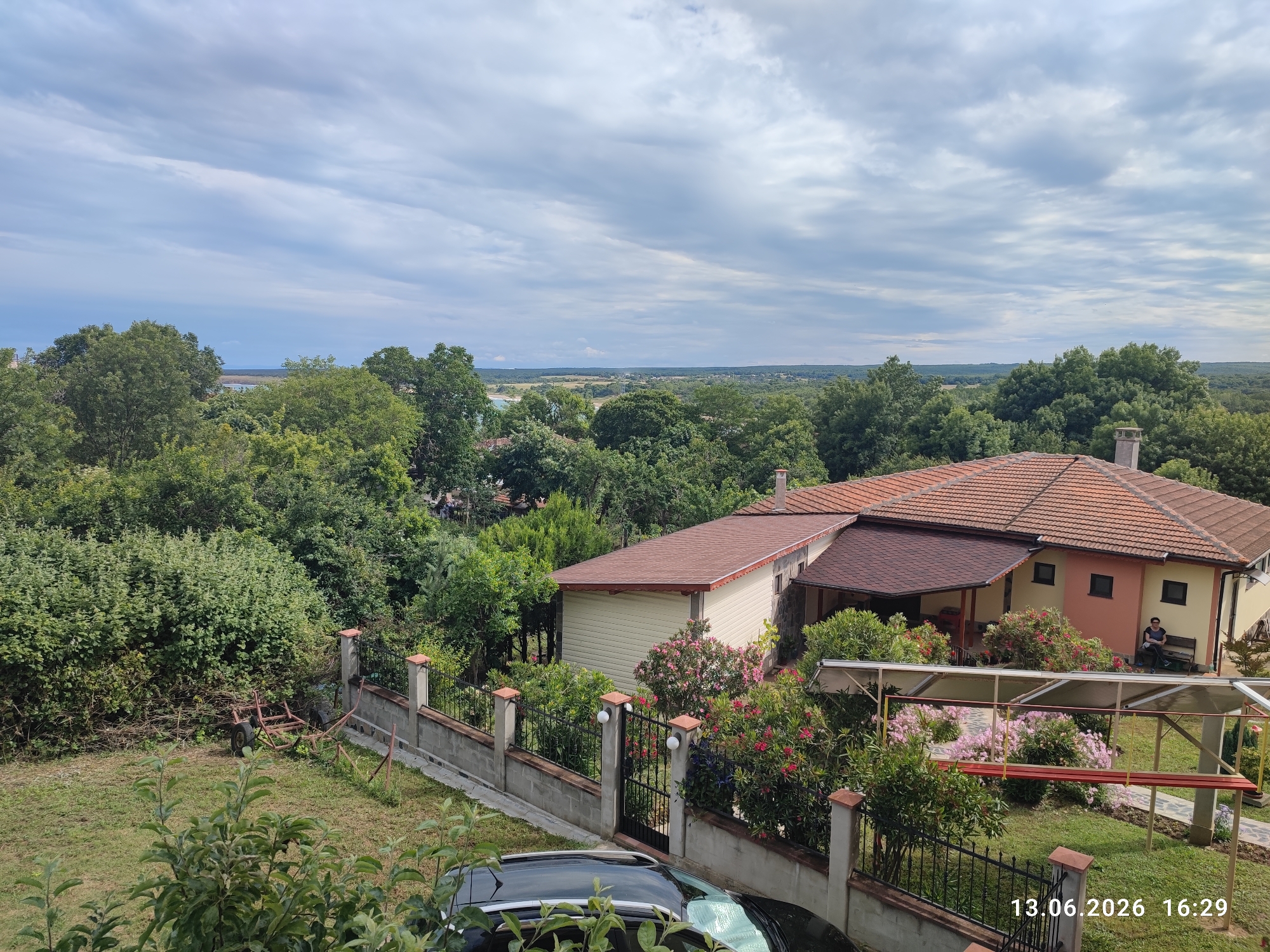 In the city of Rezovo, Bulgaria, a view overlooks a residential area with lush greenery and a partially cloudy sky. In the foreground, a dark-colored car is parked on a grassy area. A concrete and metal fence separates the yard from a property with a house and an attached garage. The house has a red-tiled roof and beige and pink walls. Pink and red flowering bushes are prominent in the garden. To the right of the house, a woman sits on a bench. In the background, a vast expanse of trees extends to the horizon, where a body of water and distant land can be seen. A date stamp indicates the image was taken on 13.06.2026 at 16:29.