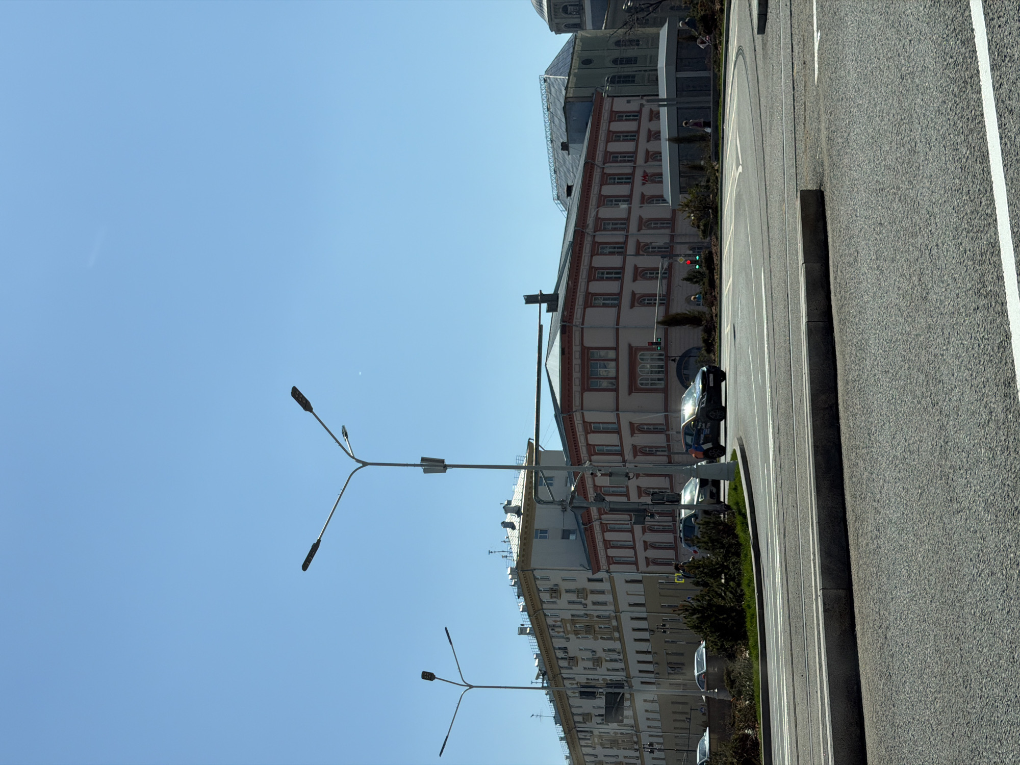 The image depicts a street scene in Moscow, Russia, characterized by a clear blue sky. The focal point is a row of buildings with a European architectural style, featuring multiple stories, ornate windows, and a mix of colors. A black car is visible parked on the side of the road. A street runs parallel to the buildings, separated by a curb and some shrubbery. Several lampposts line the street, contrasting with the blue sky. The overall impression is a typical urban scene in a European city.