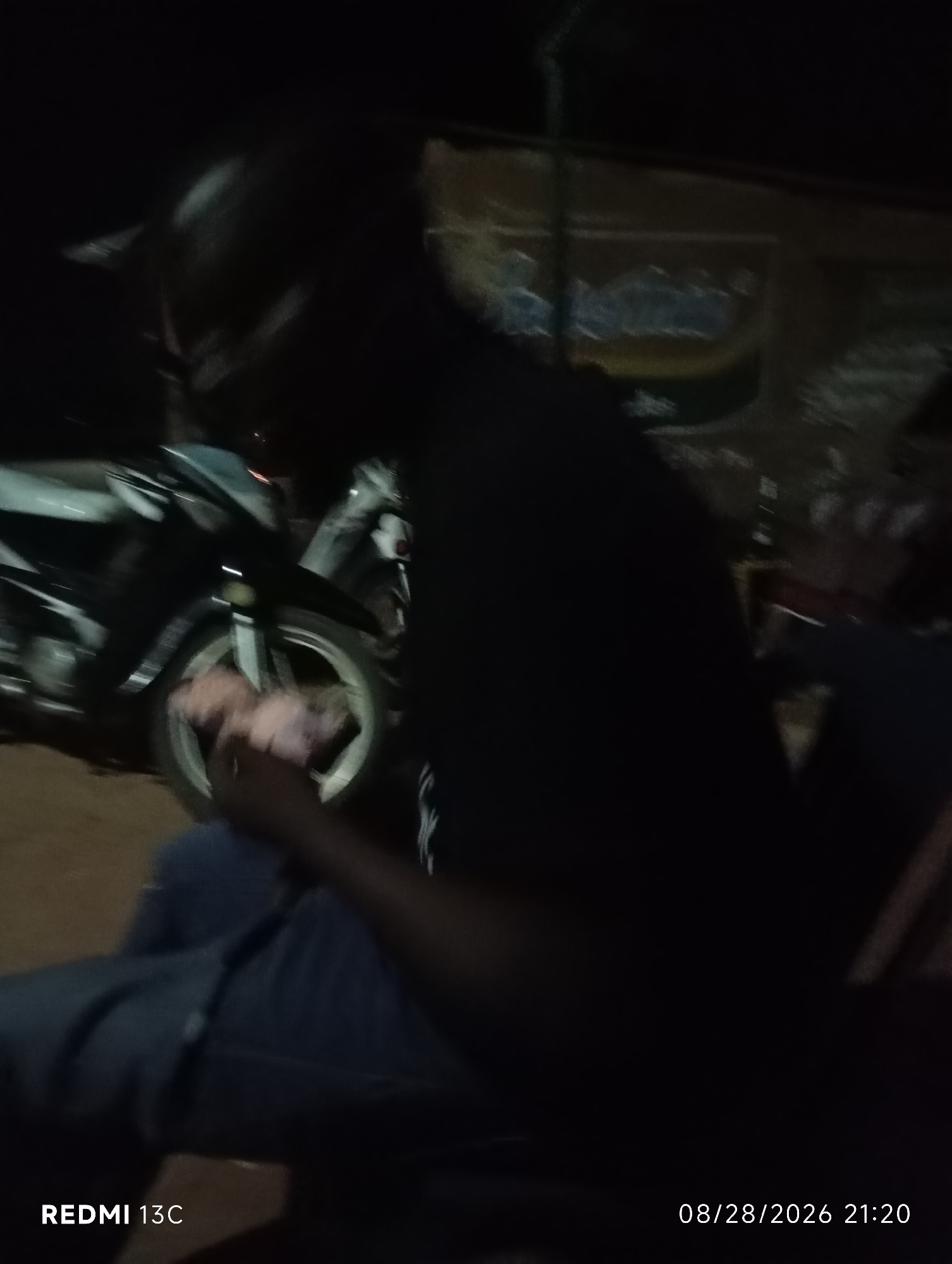 The image displays a dimly lit outdoor scene, likely at night, with a focus on a person sitting in the foreground. The person is wearing dark clothing, and their legs are crossed. To the left of the person, a motorcycle is partially visible, its front wheel and part of its body in view. The background is dark and indistinct, but appears to show some structures or foliage. The bottom right corner of the image contains a timestamp: "08/28/2026 21:20", and the bottom left corner shows the text "REDMI 13C". The overall impression is that of a casual gathering or resting moment in an urban or semi-urban environment. Based on the provided location, this scene is situated in Shendam, Nigeria.