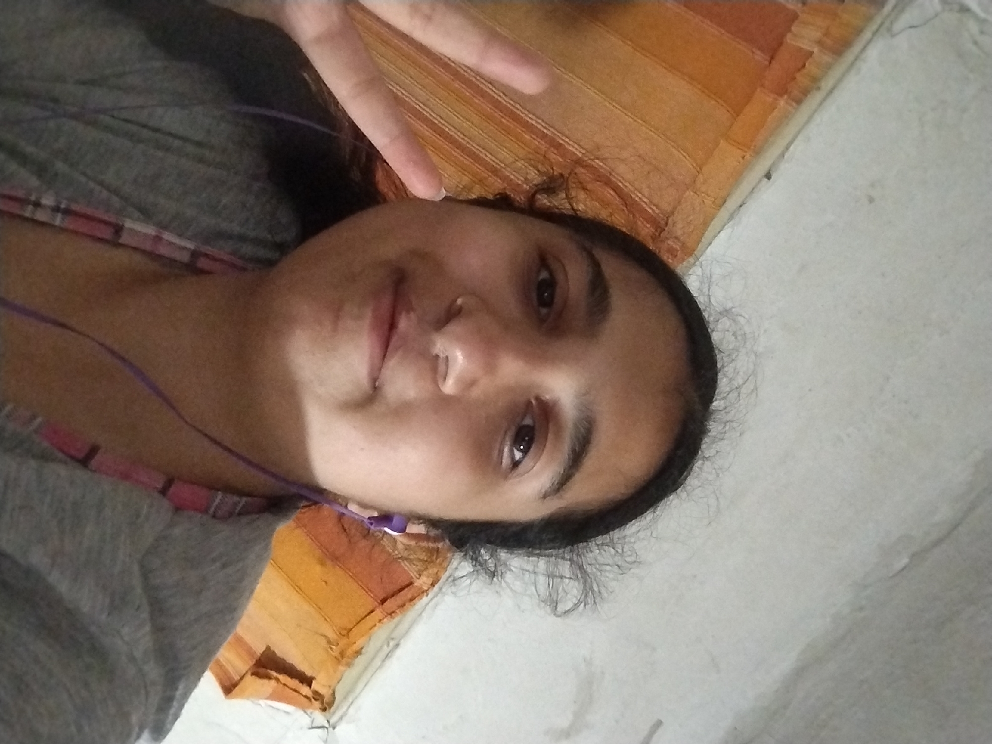 A young South Asian female is taking a selfie indoors. She has dark hair and is wearing a gray t-shirt, and purple earphones. She is smiling and holding up two fingers in a peace sign. The background consists of a wall and a patterned orange and brown fabric. The image is not taken in Plachkovtsi, Bulgaria.