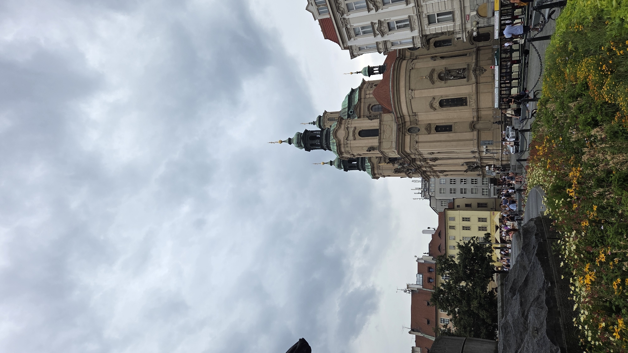 In Prague, Czechia, the St. Nicholas Church dominates the Old Town Square under a cloudy sky. The Baroque church, with its distinct green domes and ornate facade, is flanked by other historic buildings. In the foreground, a low-lying flower bed bursts with yellow and white blooms, contrasting with a large, dark, abstract sculpture to the left. A group of tourists are visible in the middle ground, some seated on benches, others milling about. A tram or tourist train is also present, parked along a chain-link barrier. The overall atmosphere is overcast and bustling with pedestrian activity.