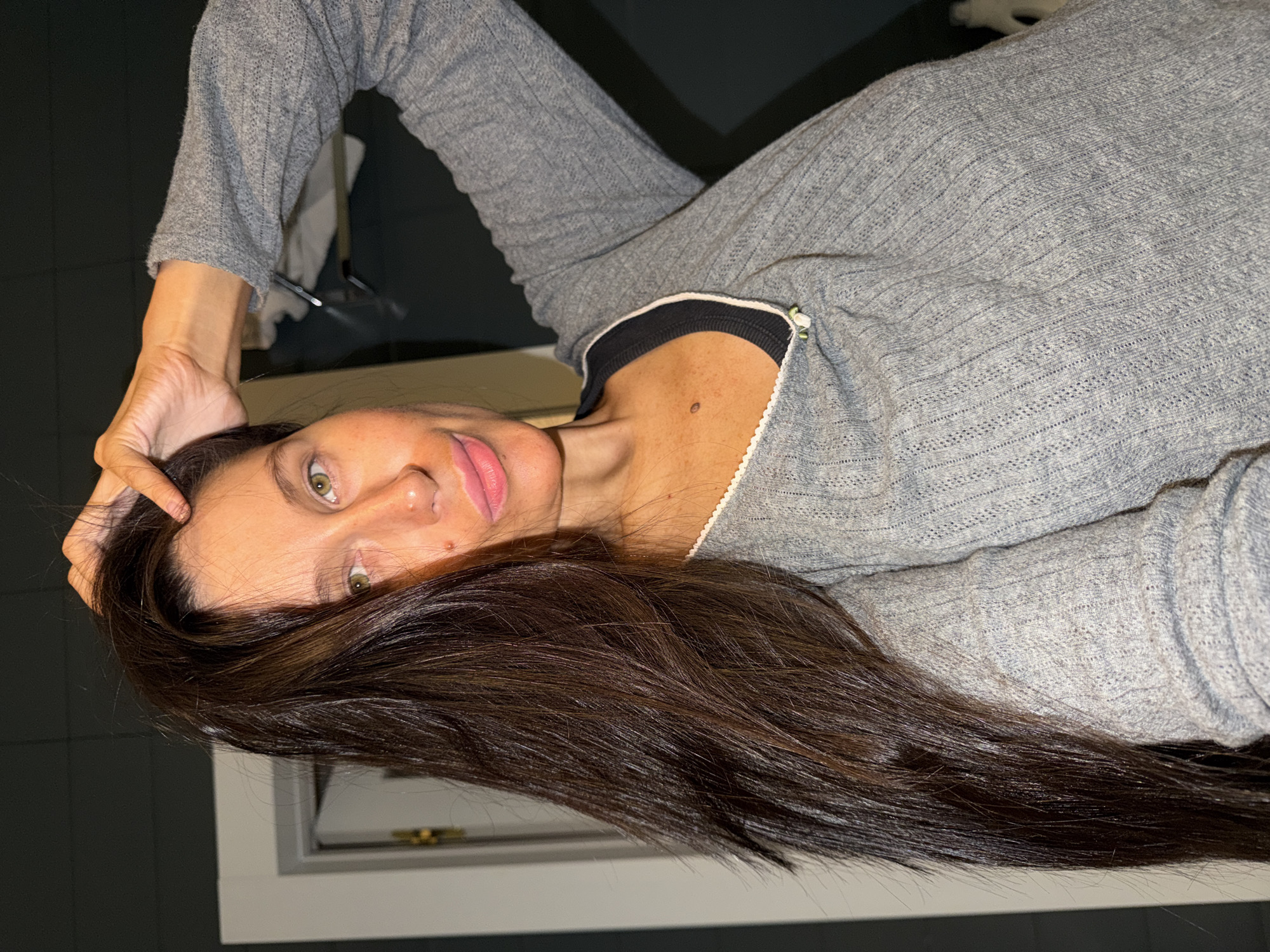 A woman with long, dark brown hair is posing for a selfie indoors. She is wearing a gray, ribbed long-sleeved shirt with a black underlayer and white trim. She has fair skin, green eyes, and a mole on her neck. Her right hand is resting on her head, pulling her hair back slightly. The background appears to be a dark gray wall and part of a window frame. There is no information in the image to confirm the location in the city of Khimki, Russia.