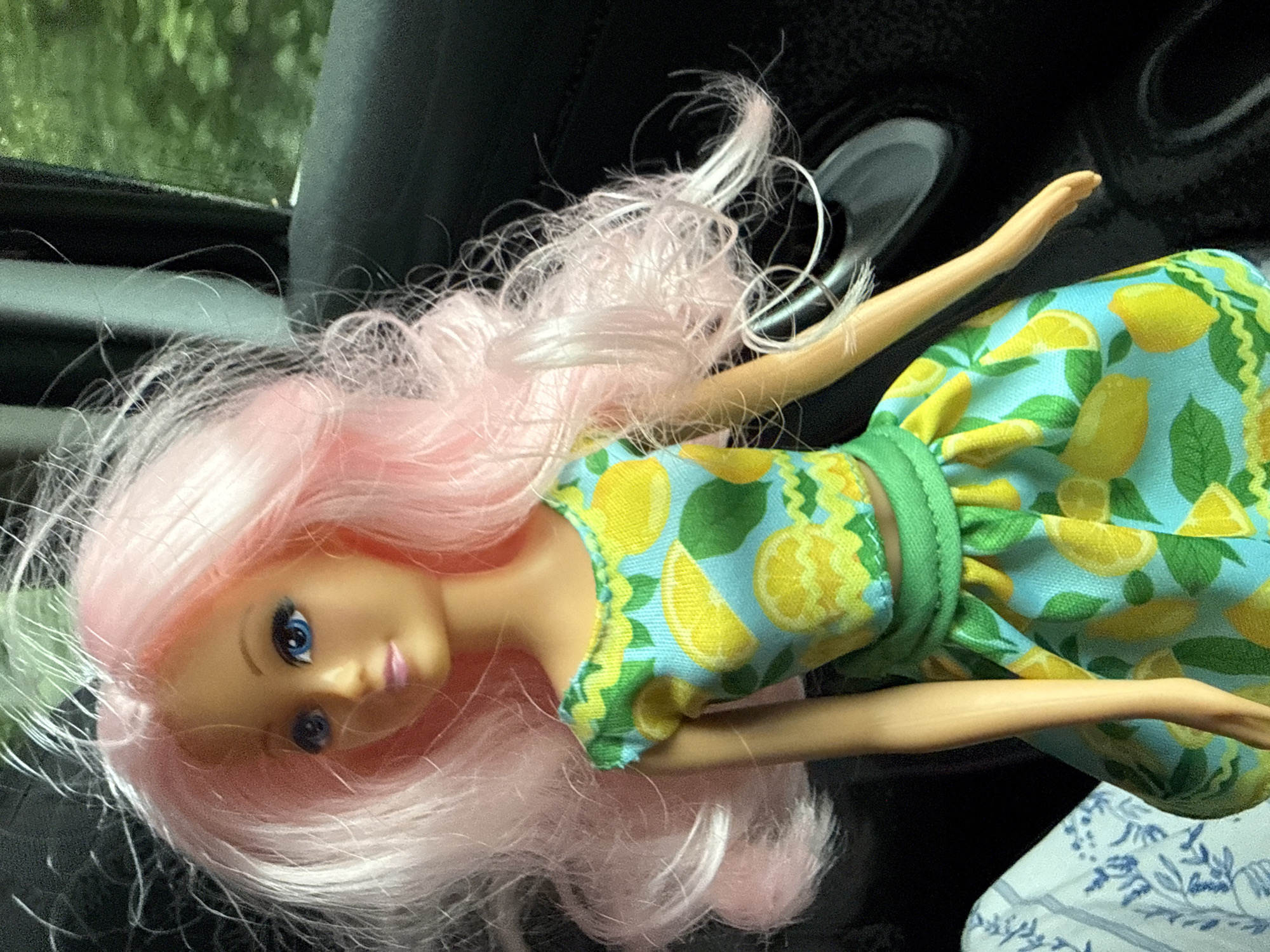 A Barbie doll with long, wavy pink hair is shown from the chest up, sitting in the passenger seat of a car. The doll is wearing a two-piece outfit: a short-sleeved crop top and a skirt, both with a blue background and a pattern of yellow lemons and green leaves. The doll's head is turned to its left, and its eyes are open and looking forward. The background of the image shows the interior of the car, including a dark gray seat, a car door, and a window with a blurry green view of trees outside. The doll appears to be sitting near the center of the frame, with its body slightly angled. The lighting suggests it is daytime, possibly with sunlight filtering through the car window. The location in the city of Weidenhausen, Germany is not discernible from the image itself.