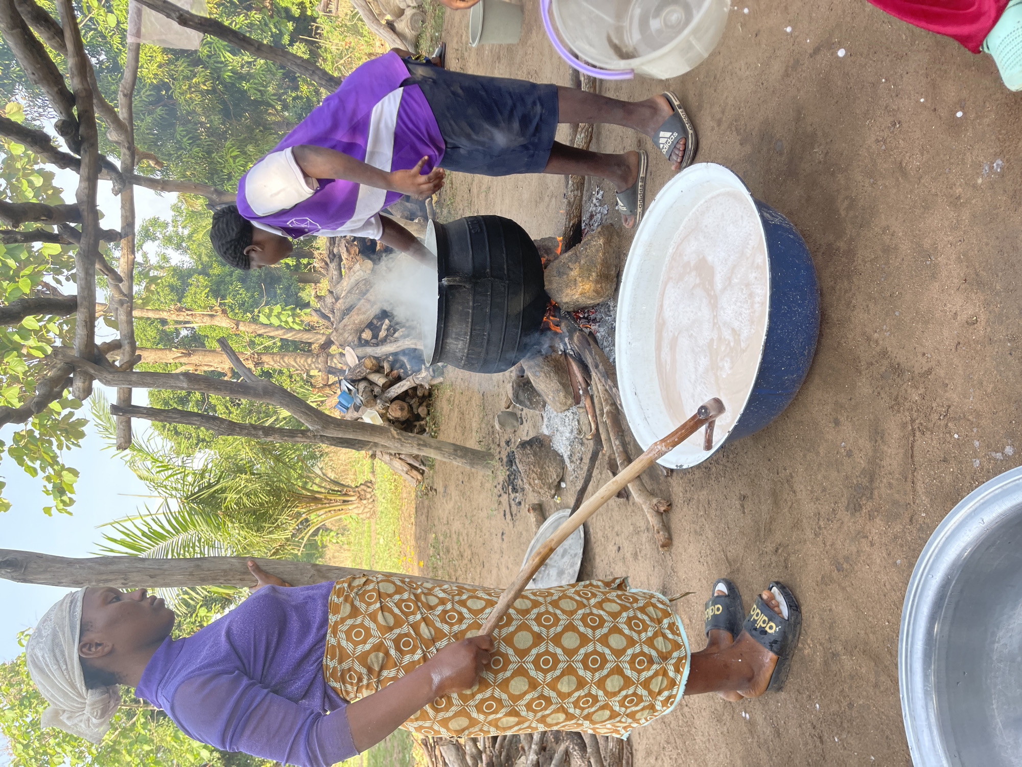 The image depicts an outdoor cooking scene in Tabu, Nigeria, with two individuals engaged in preparing food or drink using large pots over open fires on a bright day.
In the foreground, a woman, wearing a purple long-sleeved top, a patterned yellow and brown skirt, and a white headscarf, is stirring a thick, creamy, foamy liquid in a large blue and white pot with a long wooden stick. Her feet are visible, wearing black slides with "adidas" text.
Adjacent to her, another person, dressed in a purple and white short-sleeved top and dark shorts, bends over a large black pot set directly over a wood fire. Steam rises from this pot, indicating active cooking. This individual also wears black Adidas slides. Piles of firewood are stacked nearby, serving as fuel for the fire.
The setting is an unpaved, sandy ground, surrounded by lush green trees and foliage under a clear sky, characteristic of a rural or village environment. Simple wooden posts and structures are also visible, possibly part of a makeshift shelter or cooking area. Various plastic buckets and basins are scattered around the workspace. The scene portrays a method of communal or large-scale cooking prevalent in the region.