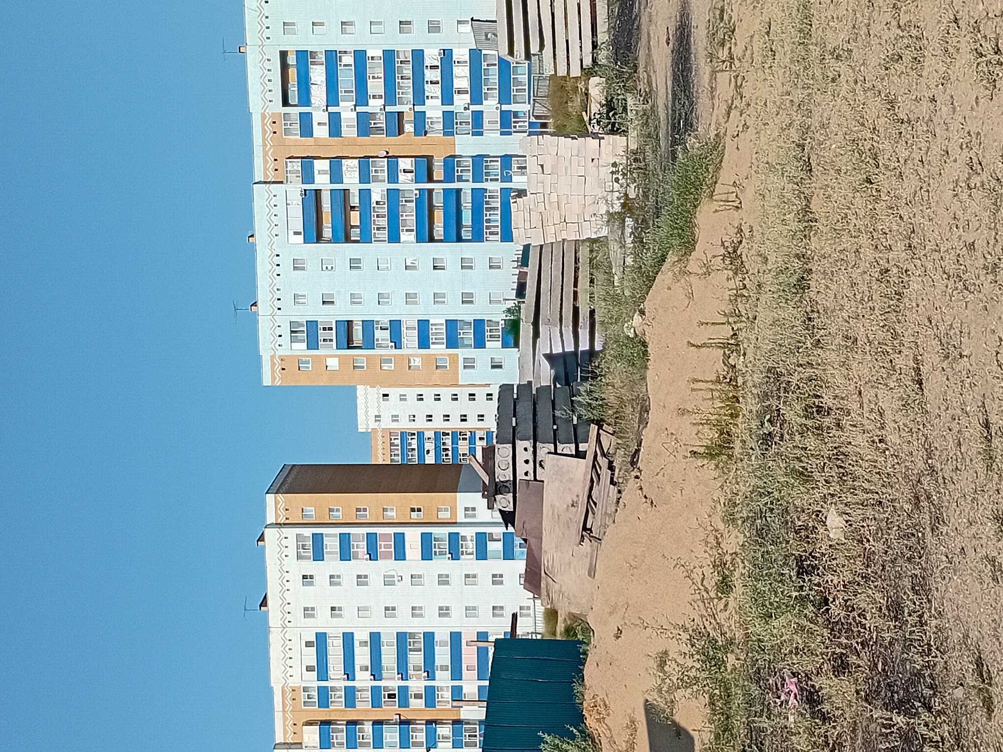 In Semey, Kazakhstan, on a clear, sunny day, the image captures a construction site in front of two multi-story apartment buildings. The buildings are light blue and tan with blue balconies and white windows. In the foreground, piles of sand, construction materials, and a green metal fence are visible. The overall scene suggests an ongoing urban development project.