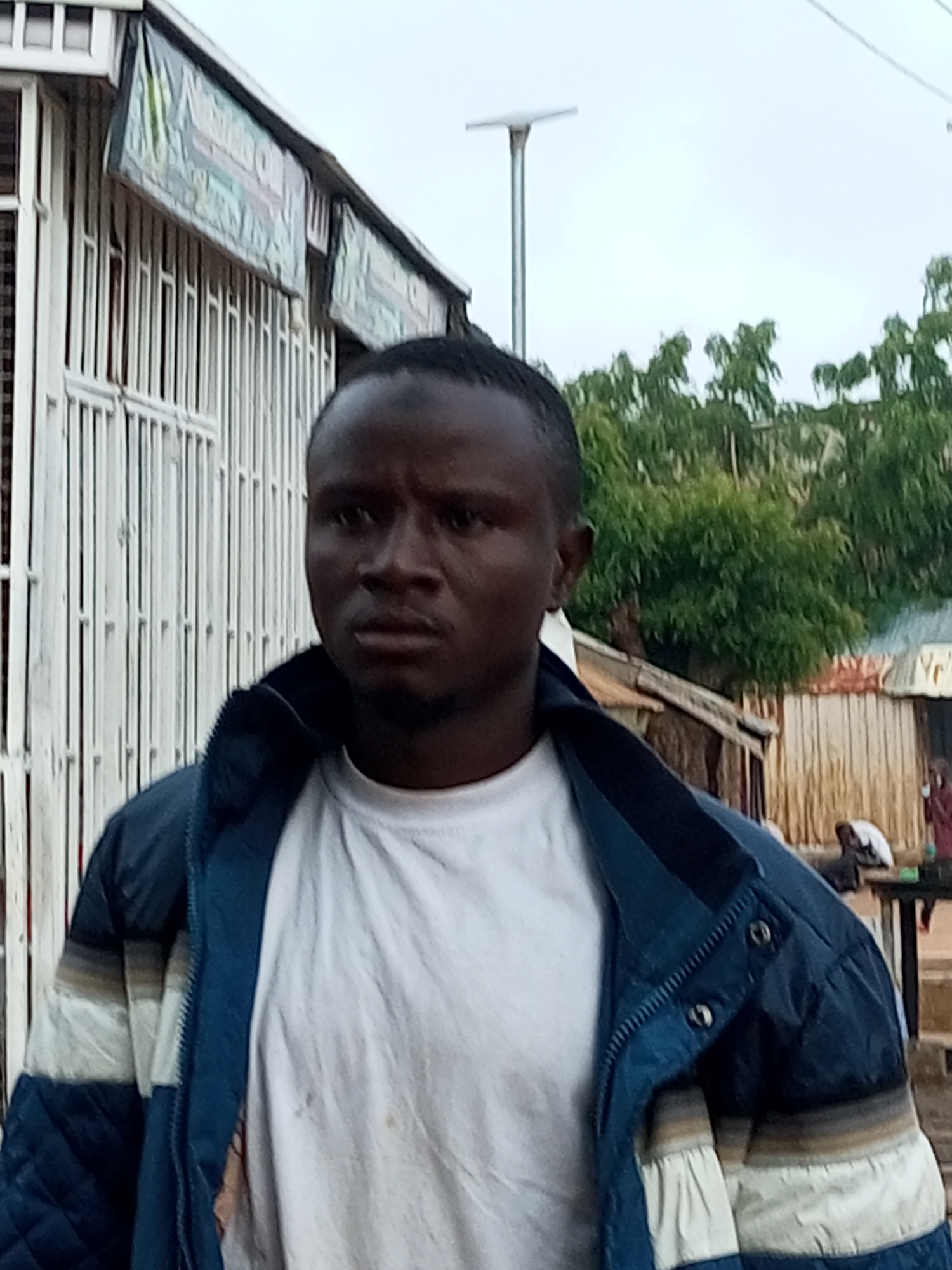 The image is a medium shot of a Black man with short, dark hair and a goatee, wearing a white t-shirt and a blue and white striped jacket. He is looking directly at the camera with a neutral expression. The man is standing in front of a white metal gate, which appears to be part of a shop. Behind the gate, there is a building with a striped awning and some lettering that is partially obscured. In the background, there are trees, a lamppost, and other buildings, suggesting an outdoor setting in a city or town. The overall impression is of a candid street portrait.