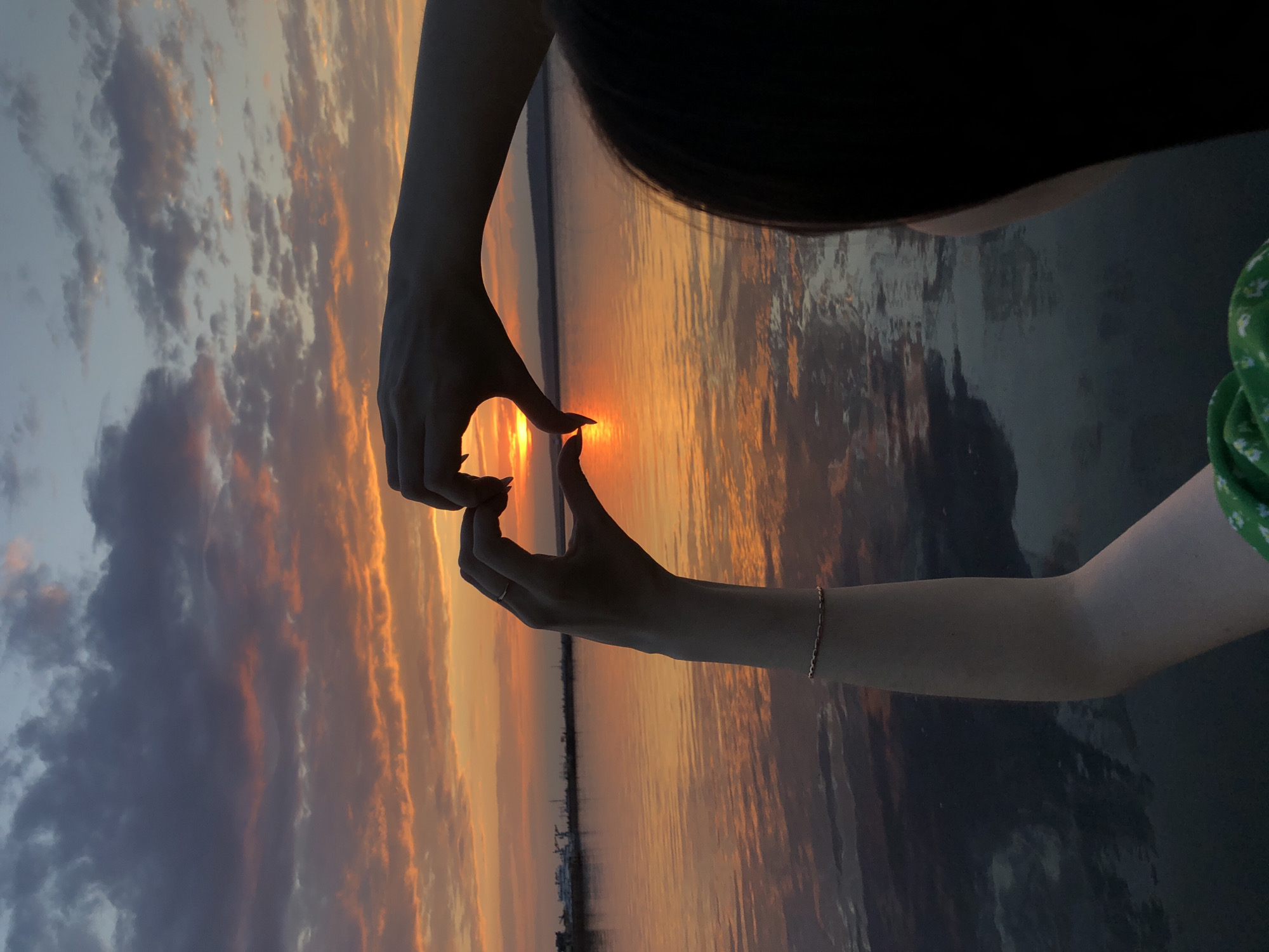 The image shows a sunset over a body of water, likely a river or lake. Two hands are positioned in the foreground, forming a heart shape around the setting sun. The sky is filled with clouds, displaying various shades of orange, pink, and gray. Reflections of the sunset and clouds are visible in the water. The angle suggests the photo may have been taken by someone on the shore or on a boat. A simple bracelet can be seen on the right wrist. Based on the location (Chaikovsky, Russia), the water body is likely the Kama River or one of the reservoirs along it.