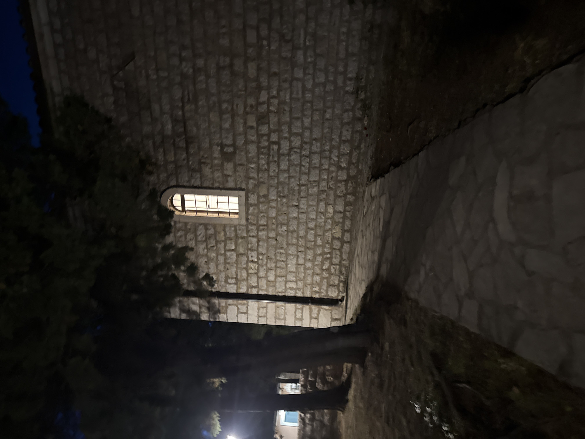 A nighttime outdoor shot shows a stone wall with a lit window. To the left of the wall, a stone path leads away from the viewer, bordered by grass and trees. The scene appears to be in an urban environment, possibly Boreti, Montenegro, given the context of the prompt. The lighting is dim, with the window providing the only significant light source.
